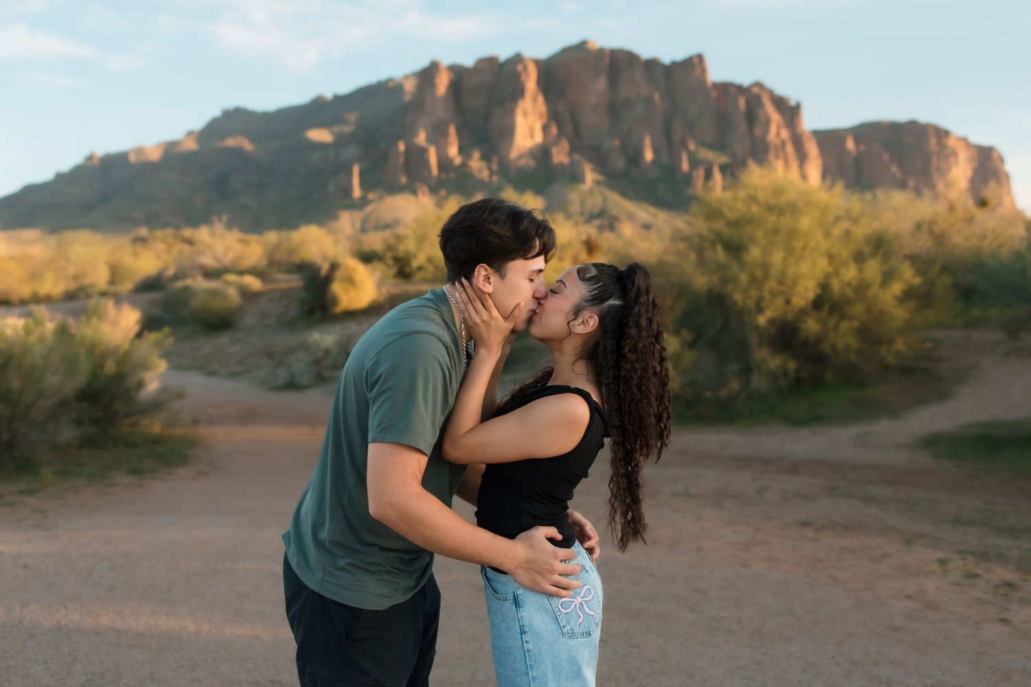 A couple shares a passionate kiss on a desert trail with dramatic mountains glowing in the sunset.