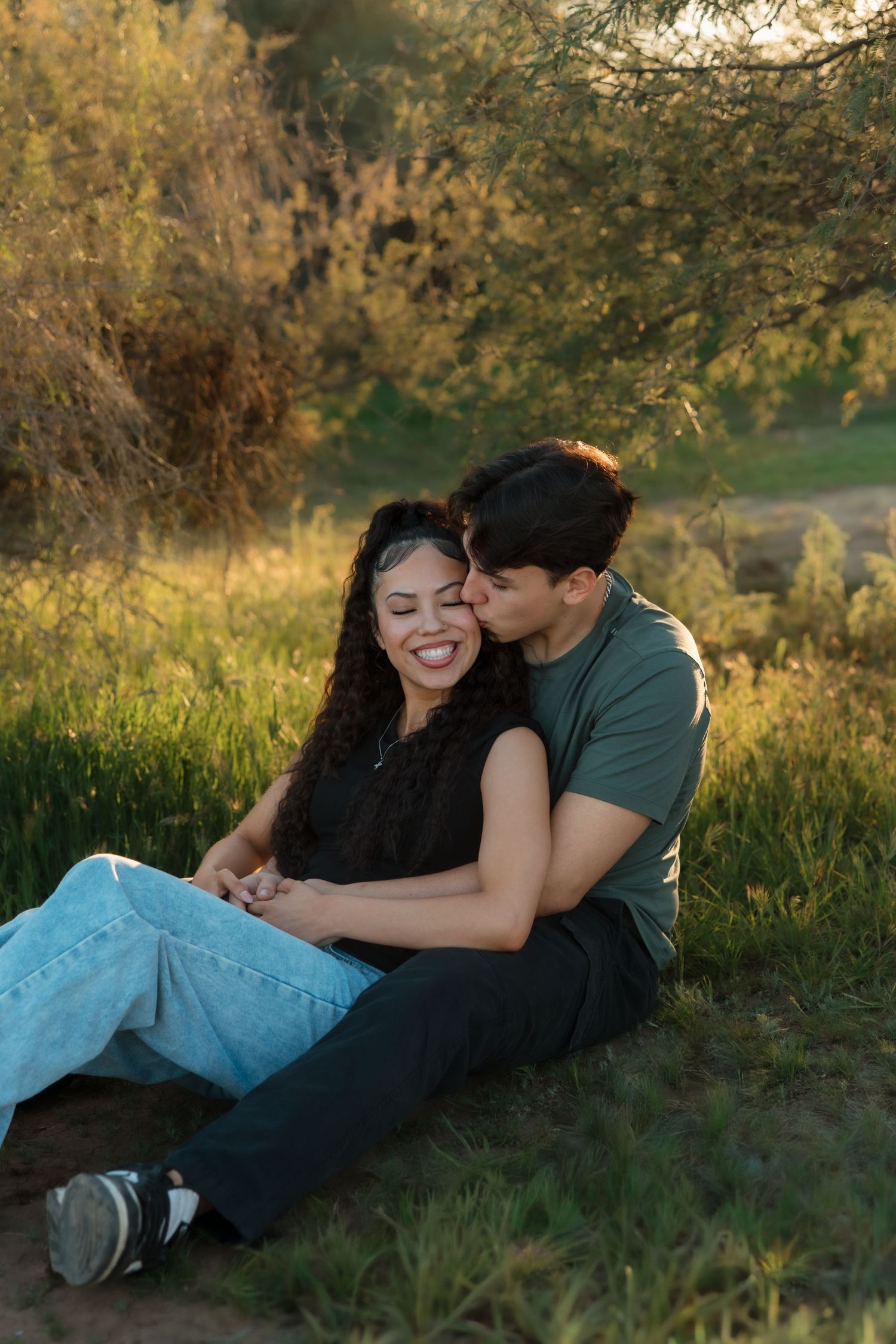 A couple sits in golden grass, the man kissing the woman's cheek as she smiles joyfully in a sunlit park.