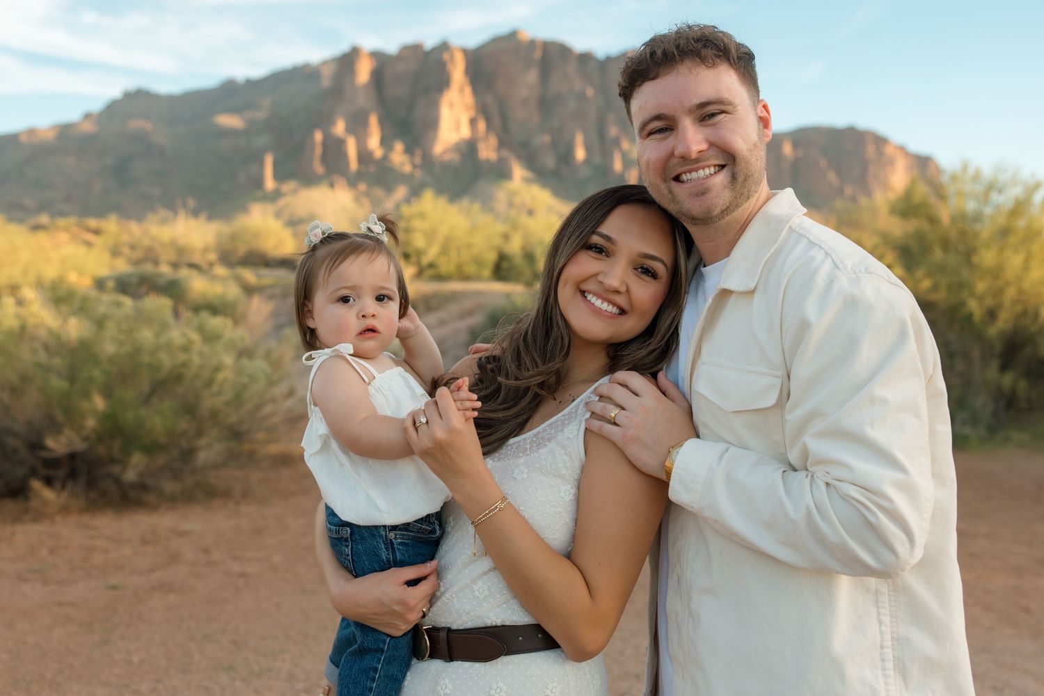 Happy family of three posing in Arizona desert with Superstition Mountains backdrop at golden hour.