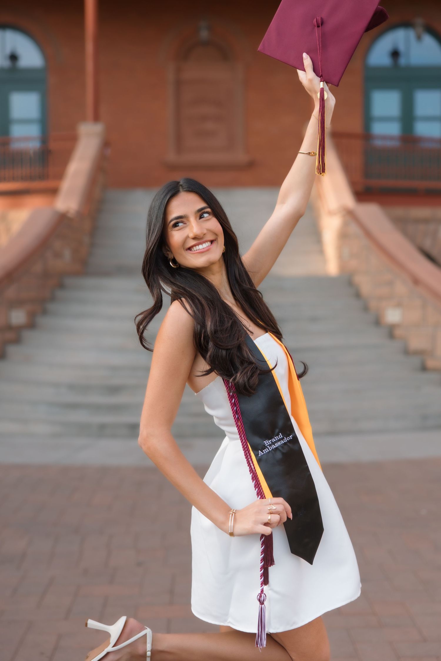 Joyful female graduate in white dress tossing maroon graduation cap in front of campus building.