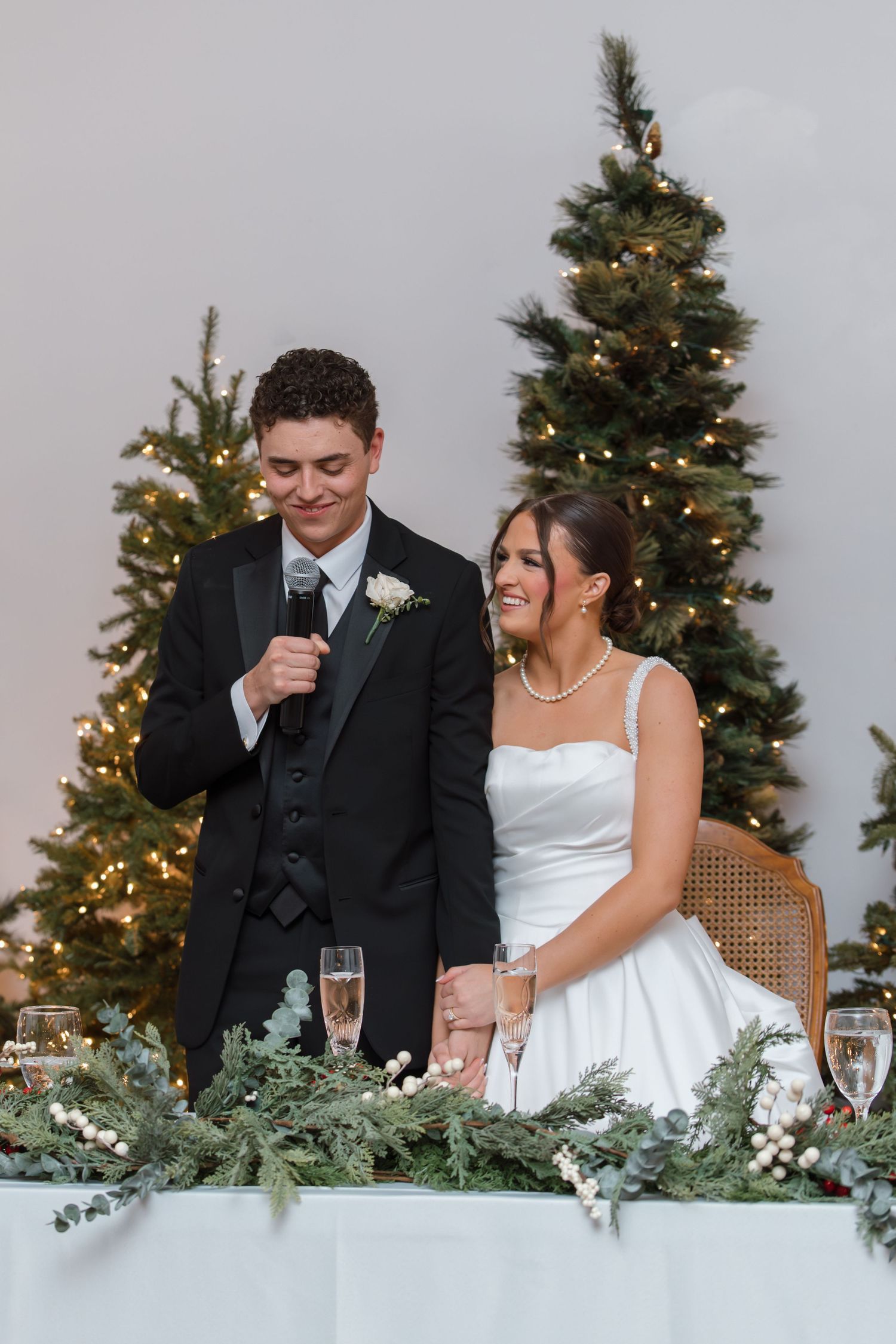 Newlywed groom speaks into microphone at sweetheart table with Christmas trees and bride smiling beside him.