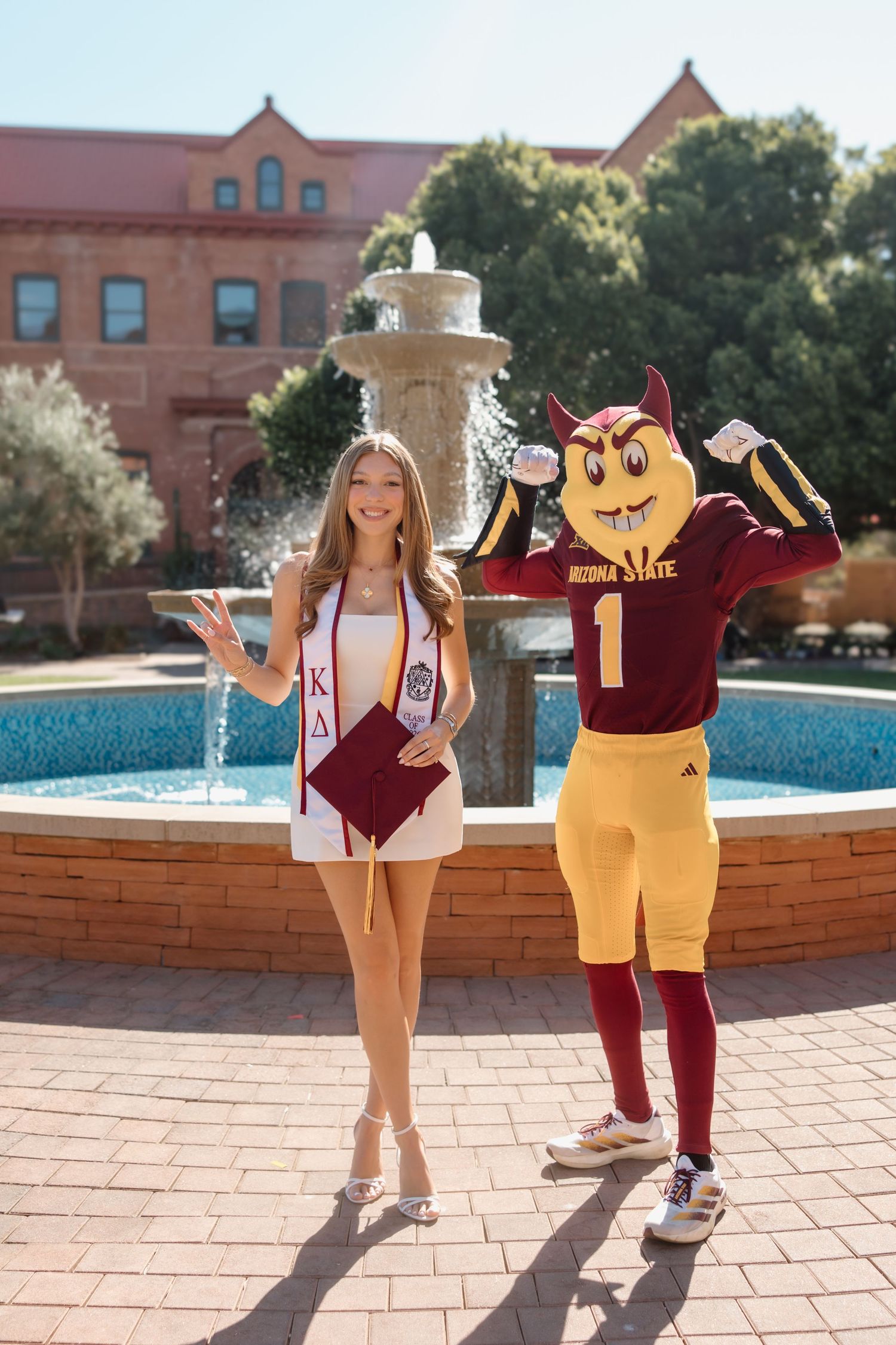 ASU graduate in Kappa Delta sash poses with Sparky mascot by campus fountain at Arizona State University.