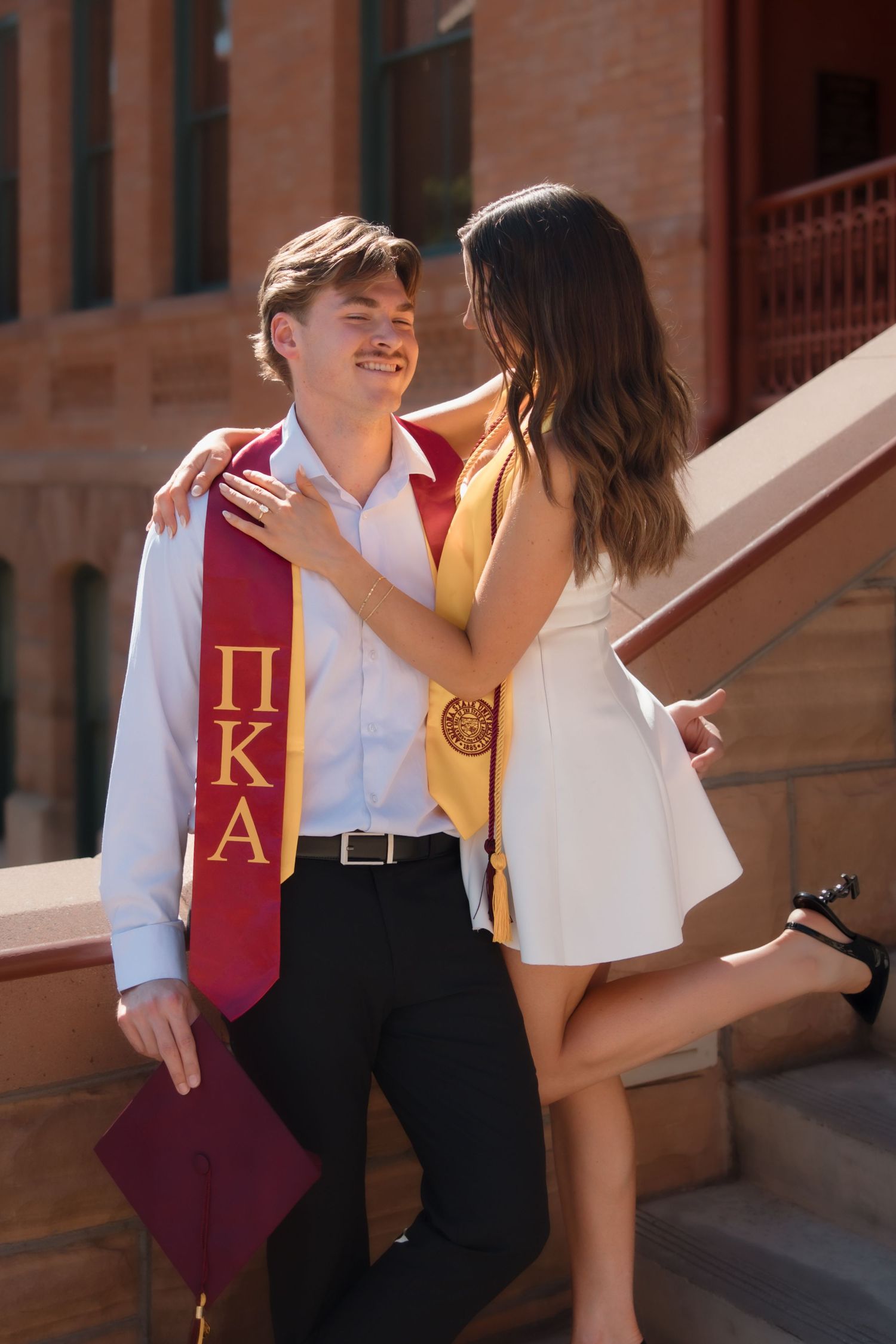 Smiling graduate wearing Pi Kappa Alpha stole embraces woman in white dress on university steps.