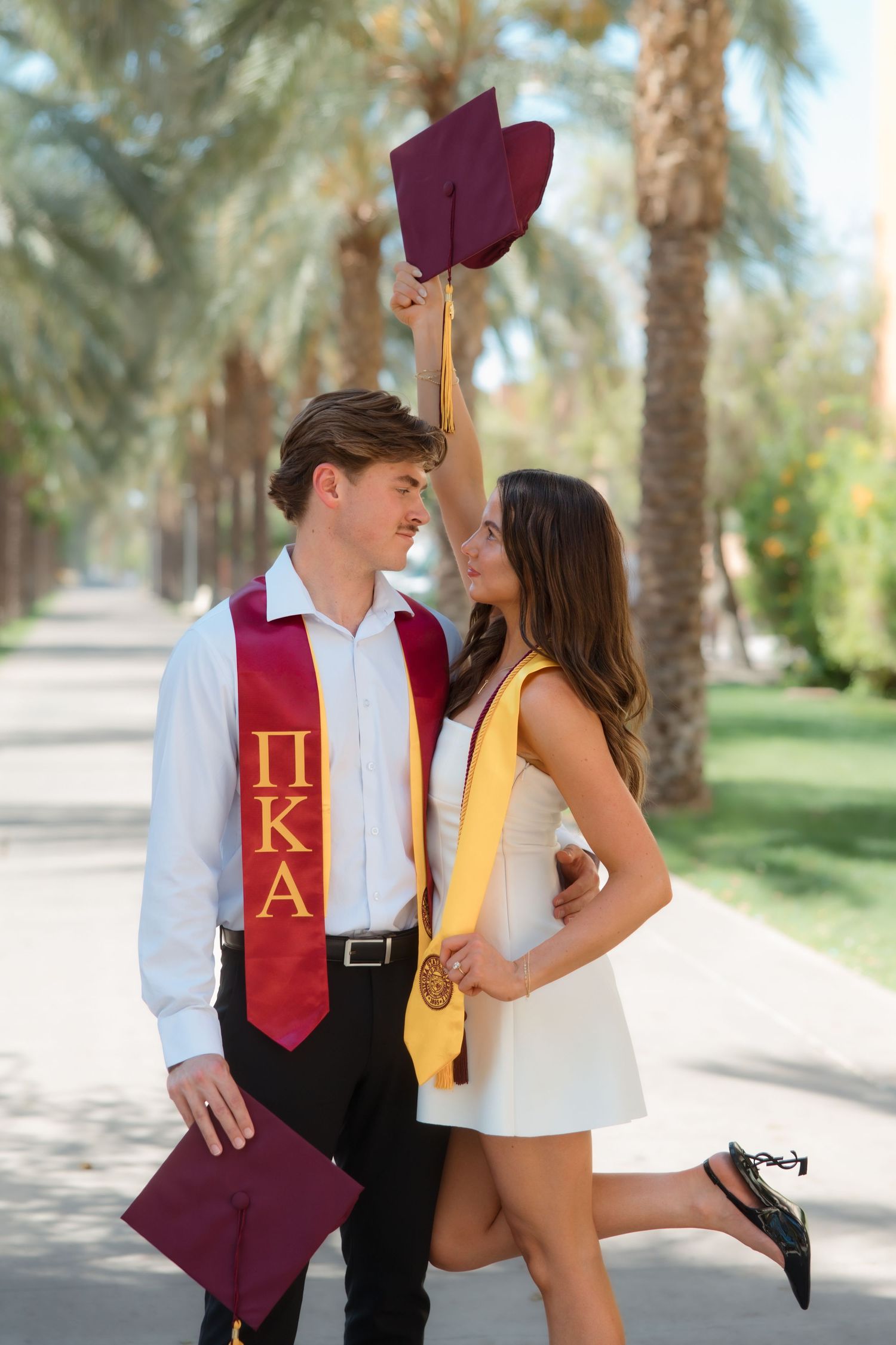 Smiling graduating couple nearly kissing, woman raising cap, on palm-lined campus path.