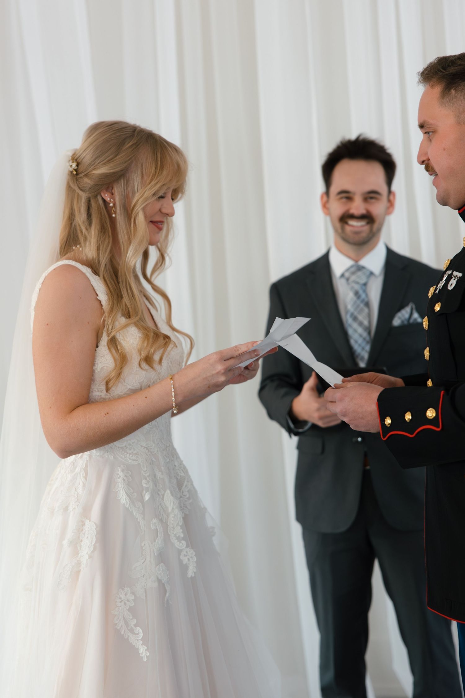 Blonde bride reads vows from paper while smiling groom in military uniform stands before officiant.