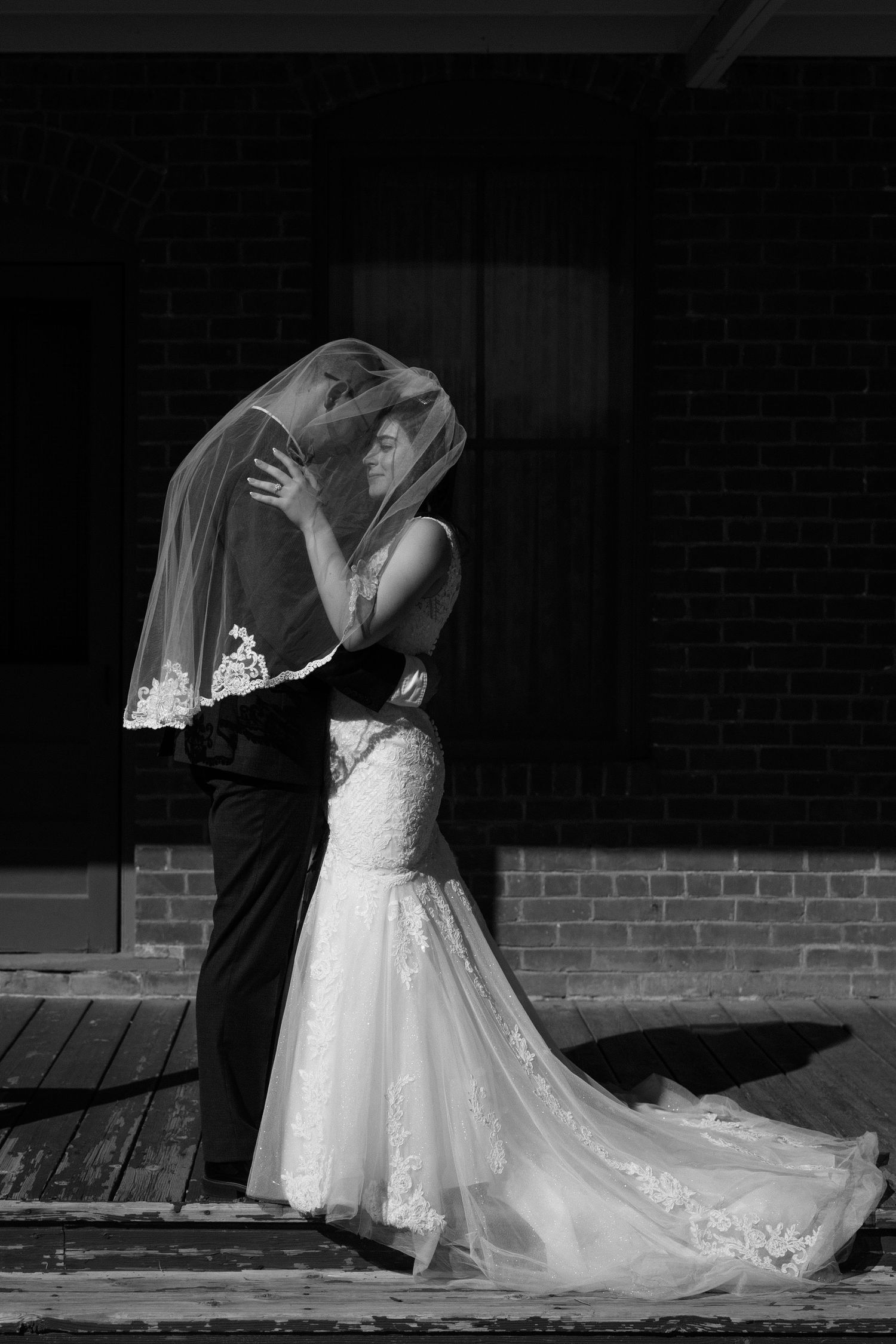 Black and white romantic photo of bride and groom kissing under flowing wedding veil outside brick building.