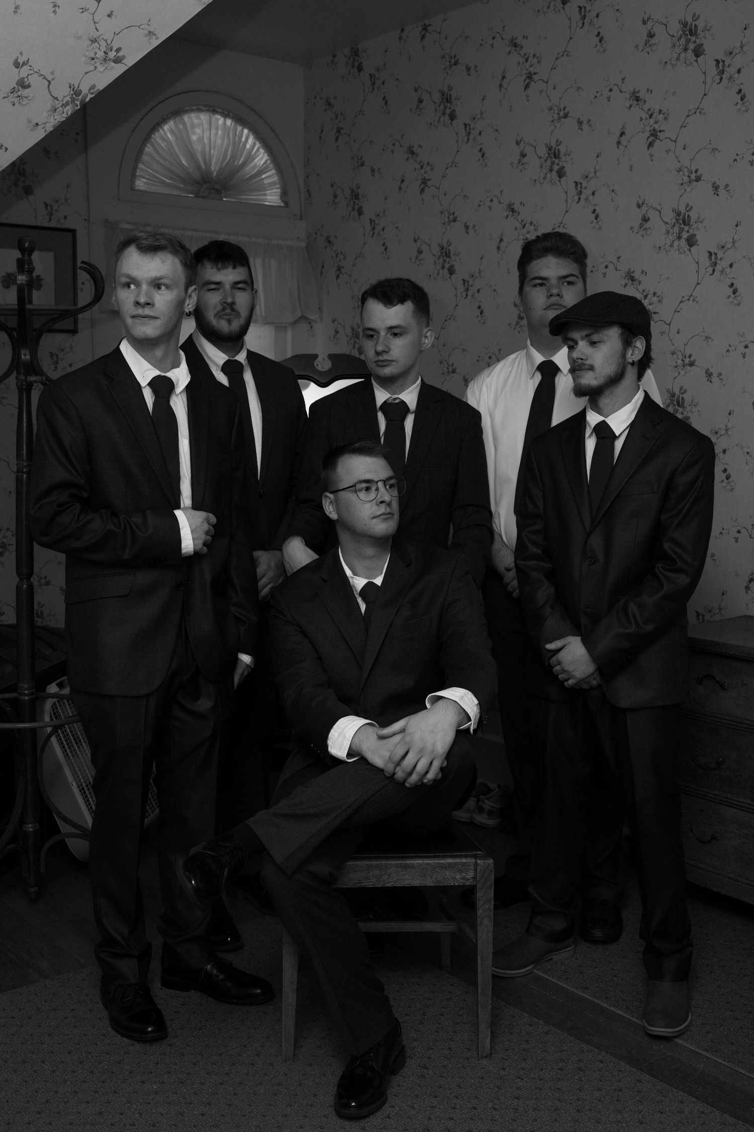 Black and white wedding photo of six groomsmen in dark suits posing in a vintage decorated room.