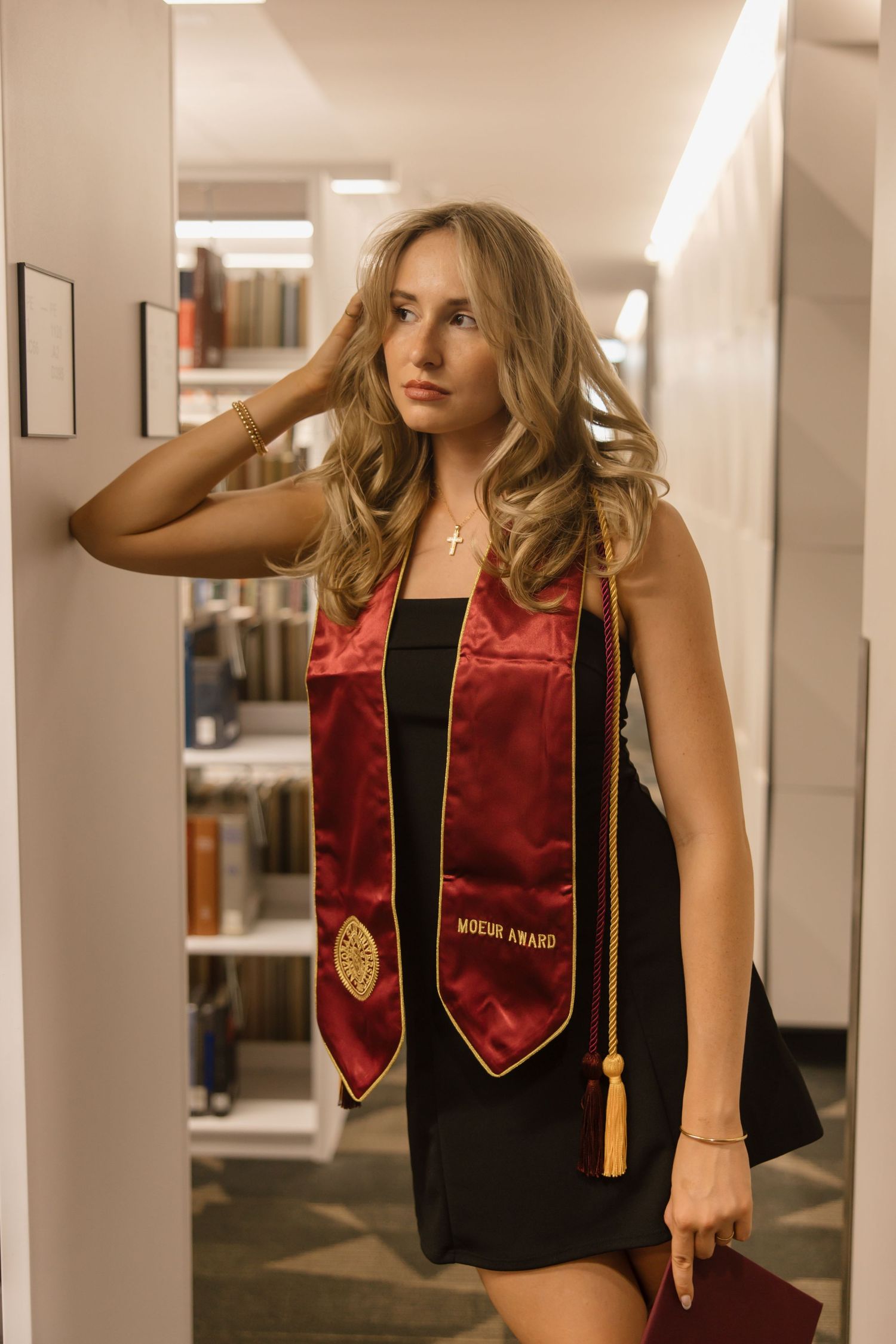 Young woman in black dress wearing red and gold graduation stole poses in a library for senior portraits.
