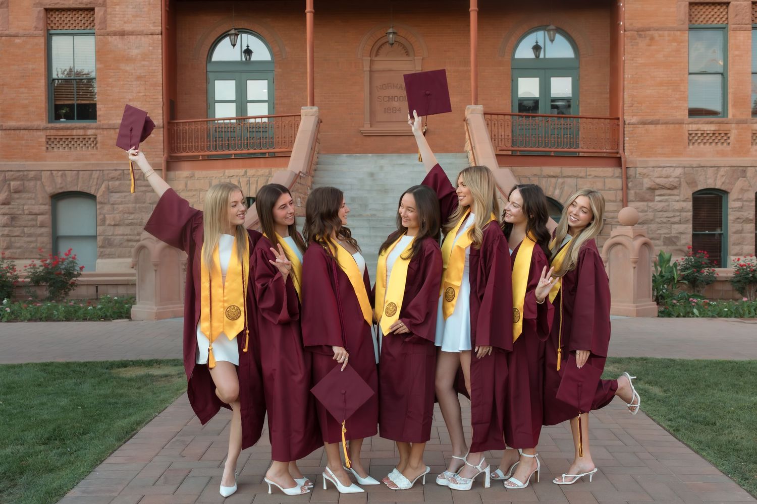 Eight female graduates in maroon and gold caps and gowns celebrate outdoors, tossing caps in front of a university building.