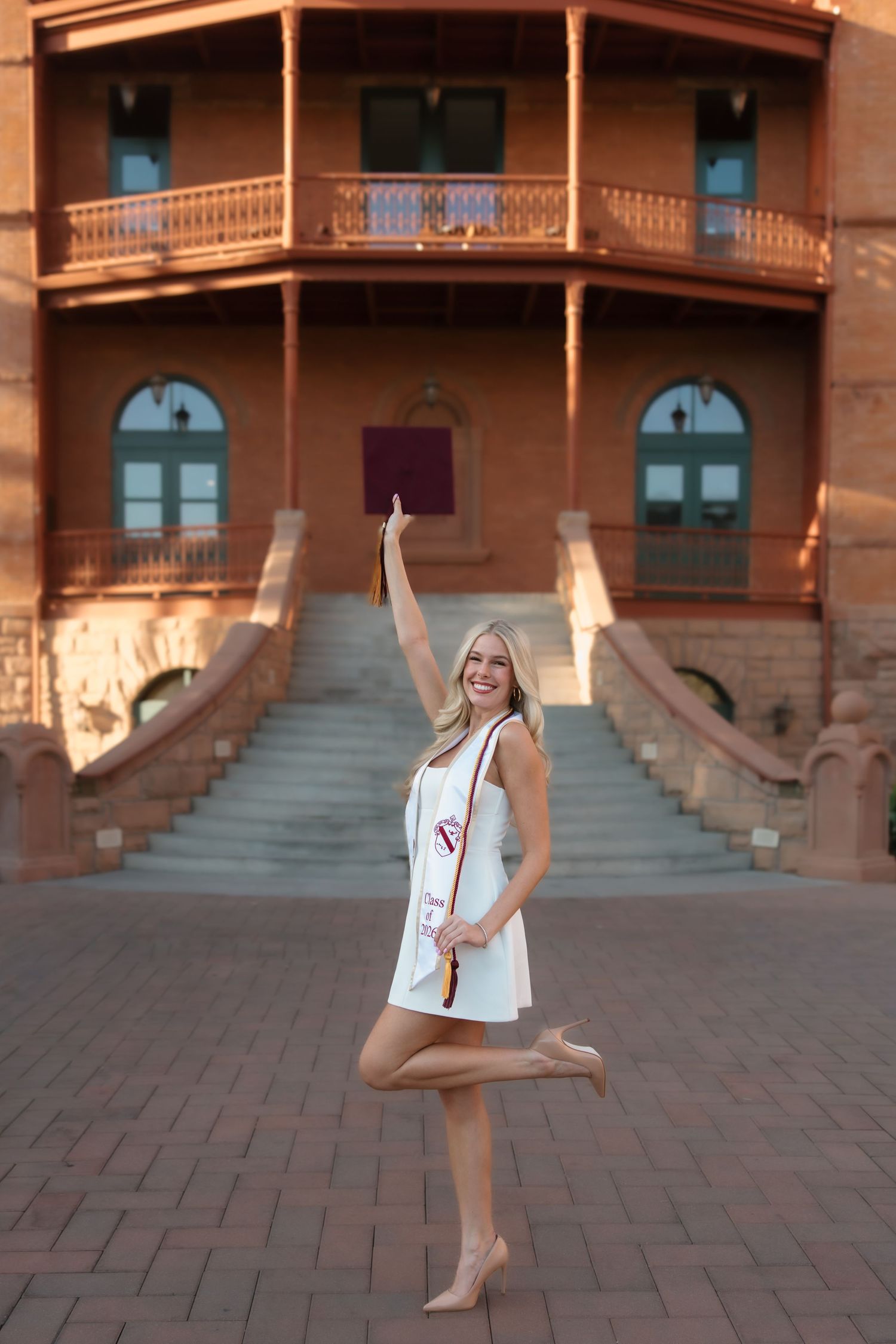Smiling blonde woman in white dress raises maroon graduation cap outside brick university building with stairs.
