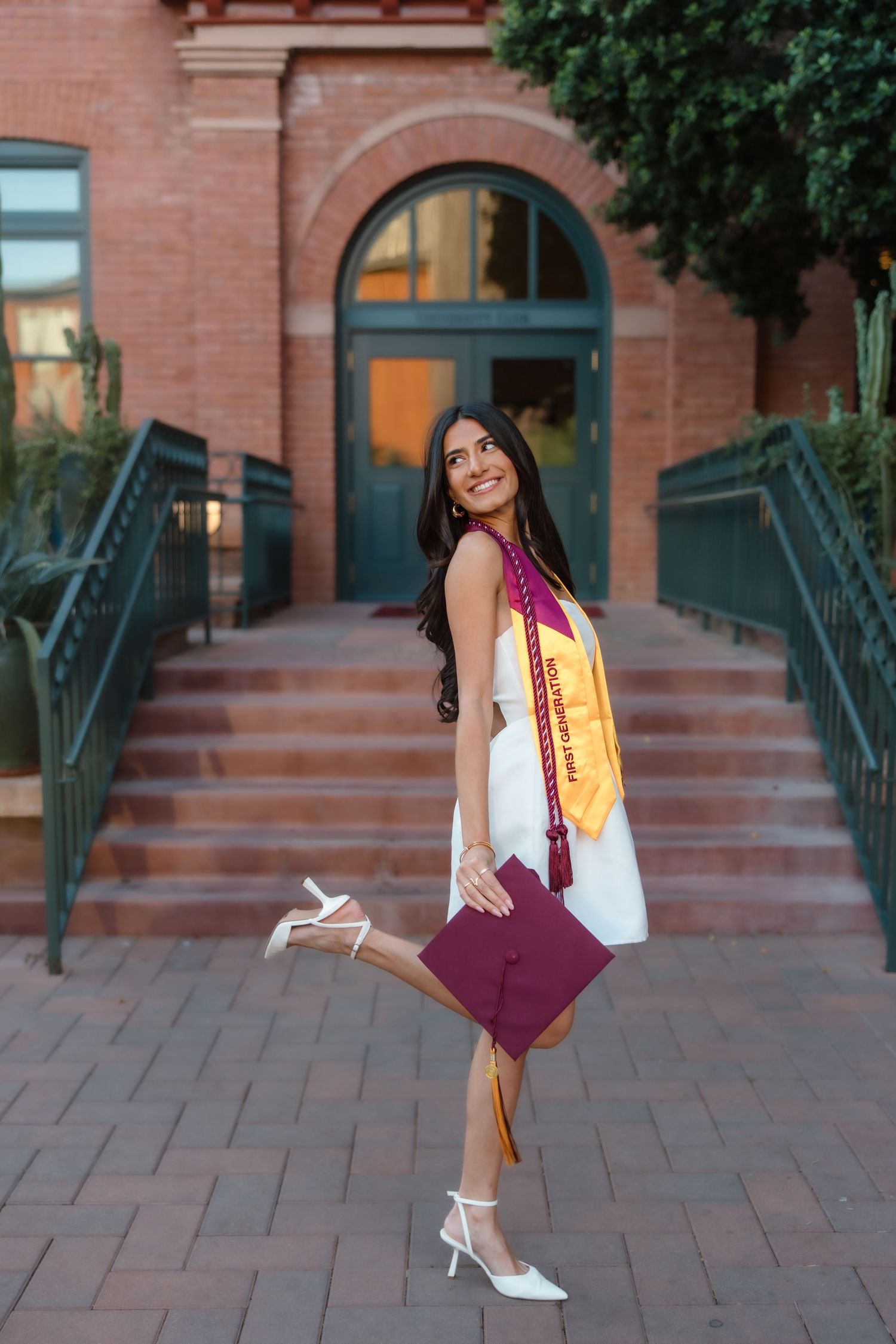 Smiling graduate in white dress holds maroon cap, wearing gold sash on brick university steps.