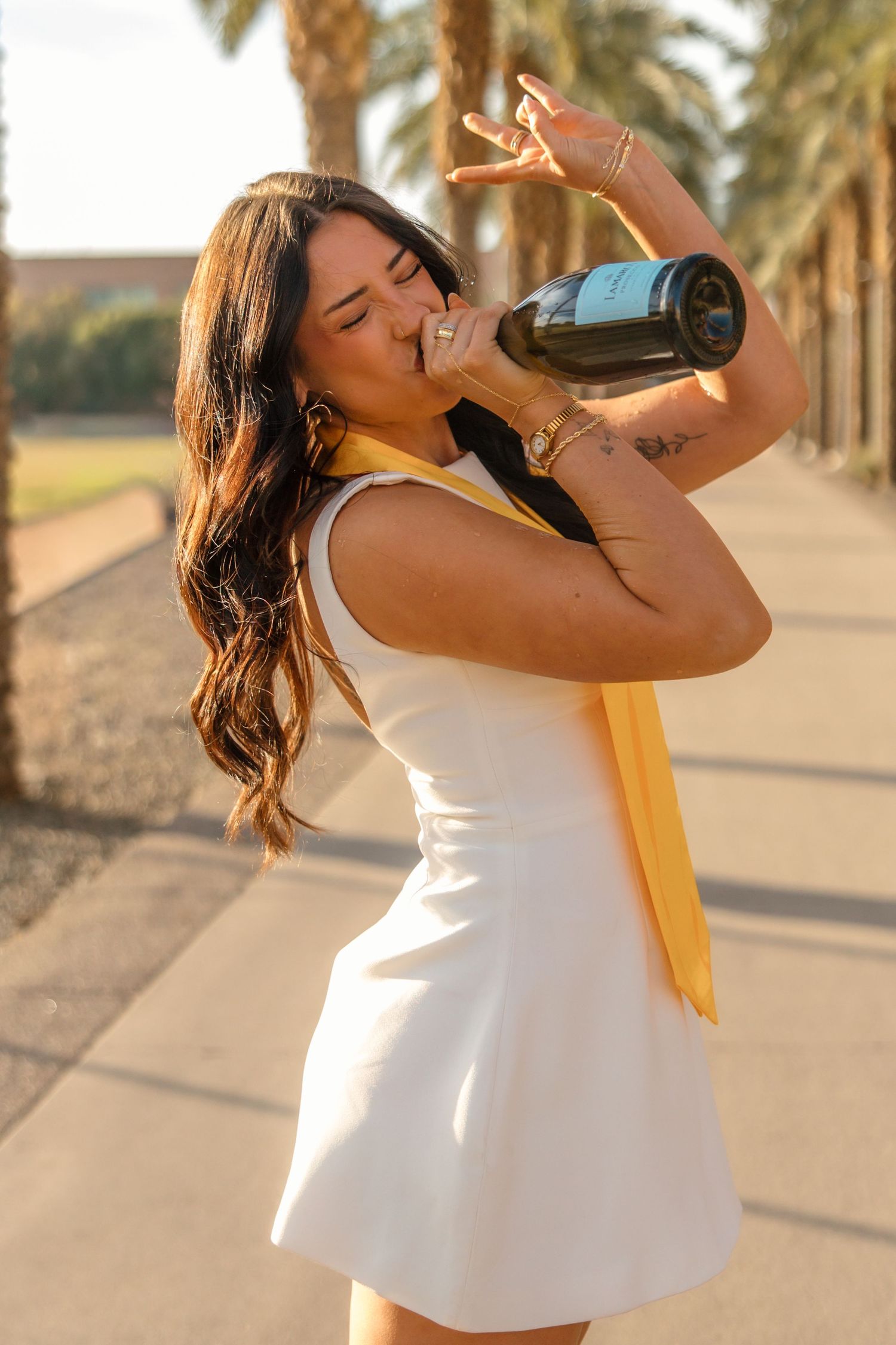 Young woman in white dress drinking champagne bottle outdoors, palm trees background, golden hour lighting.