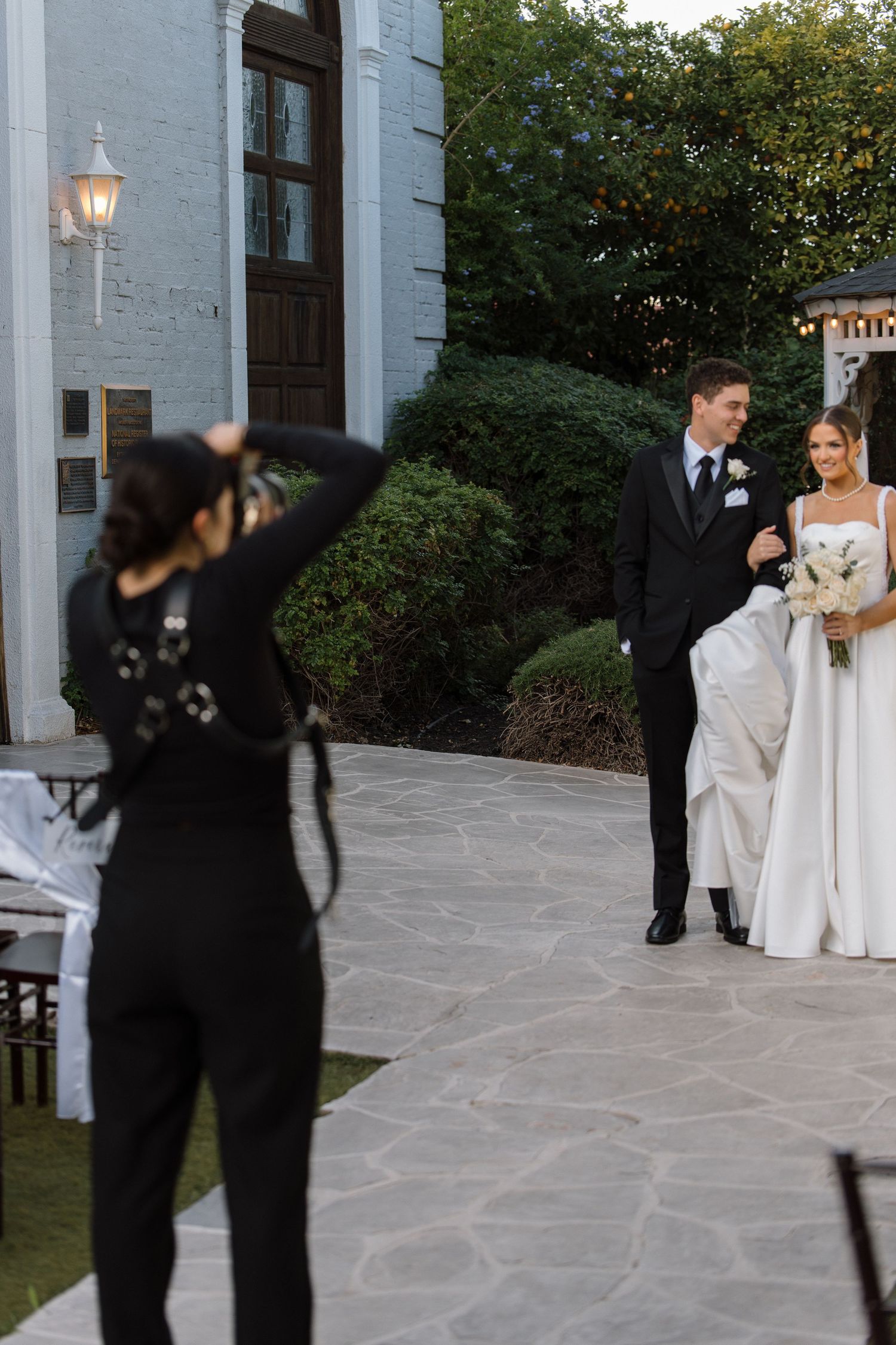 Wedding photographer captures smiling bride and groom walking on stone patio outside elegant venue.