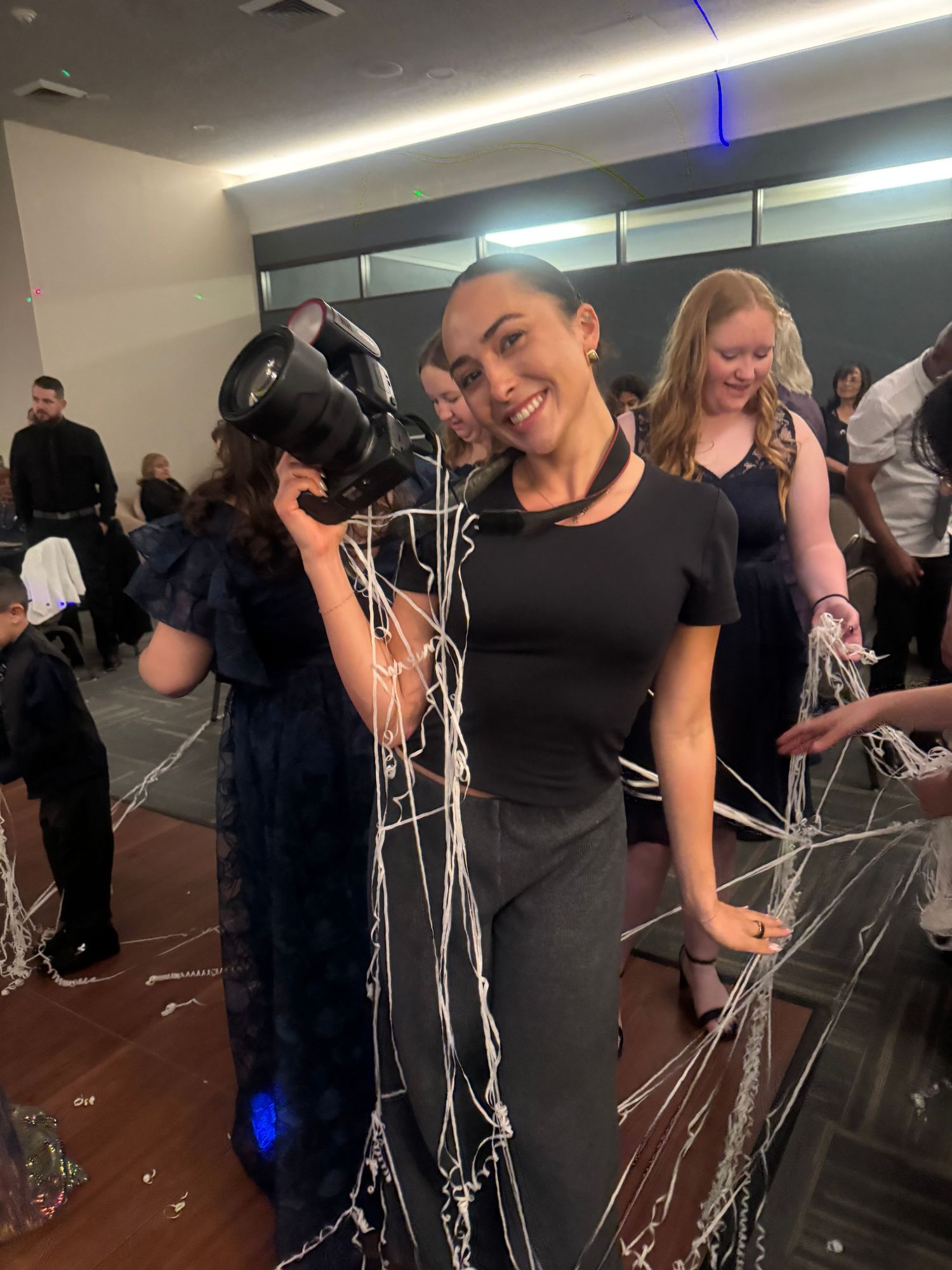 Smiling female photographer holds camera at festive indoor event with streamers falling around her.