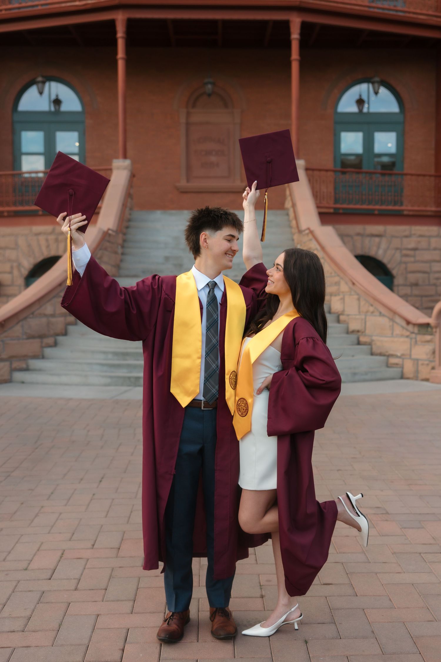 Two graduates in maroon robes and yellow stoles raising caps in front of a brick building.