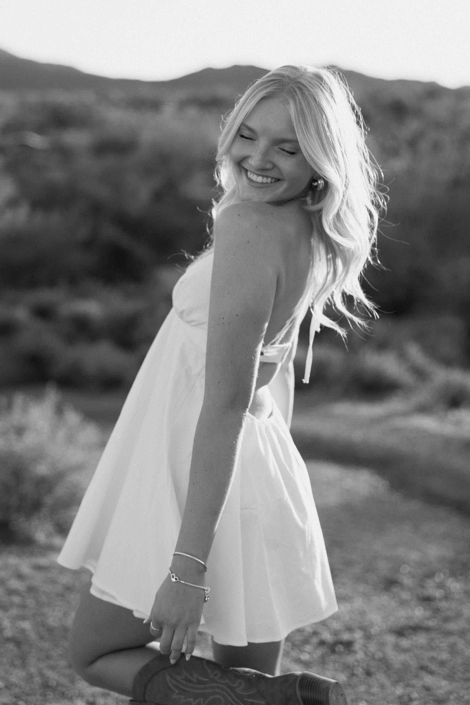 Black and white photo of a smiling blonde woman in a white dress posing outdoors in a desert landscape.
