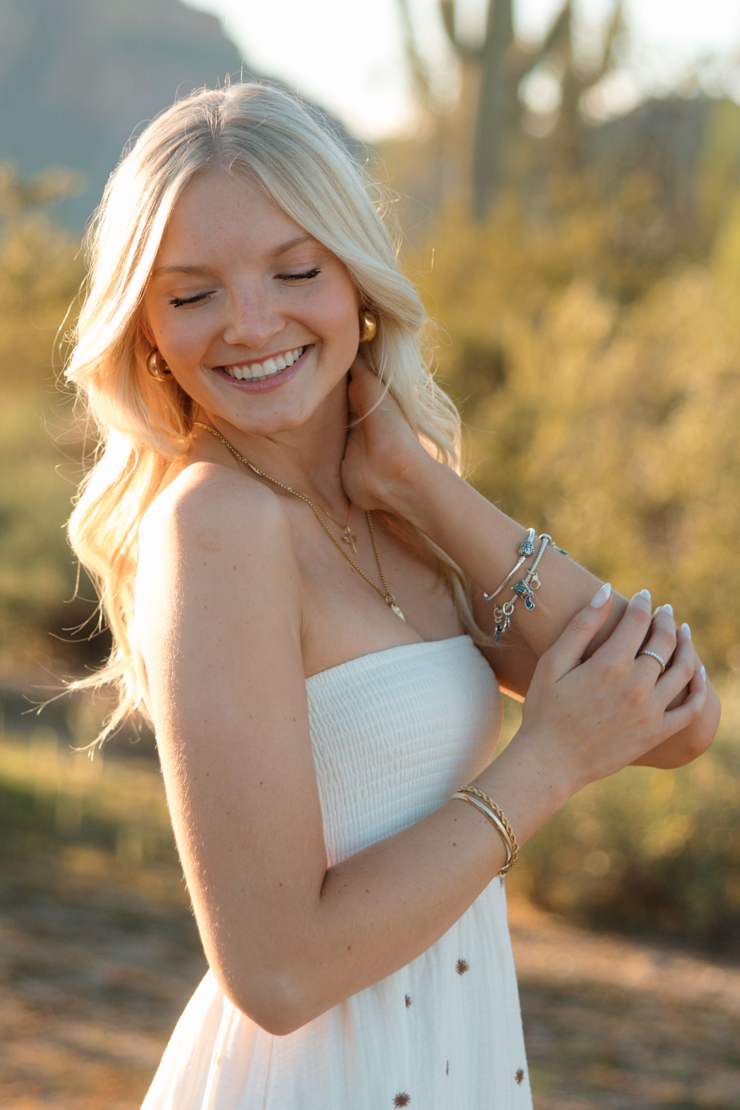 Smiling blonde woman in white strapless dress with gold jewelry, posing outdoors in desert landscape.