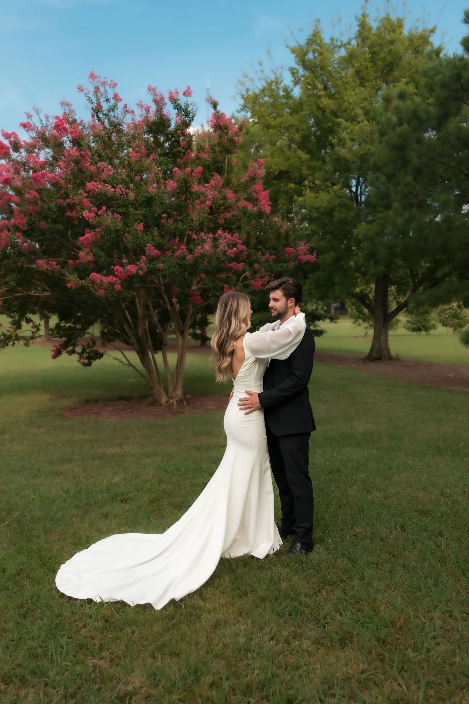 Bride and groom embracing in a lush green park with blooming pink crepe myrtle tree backdrop.