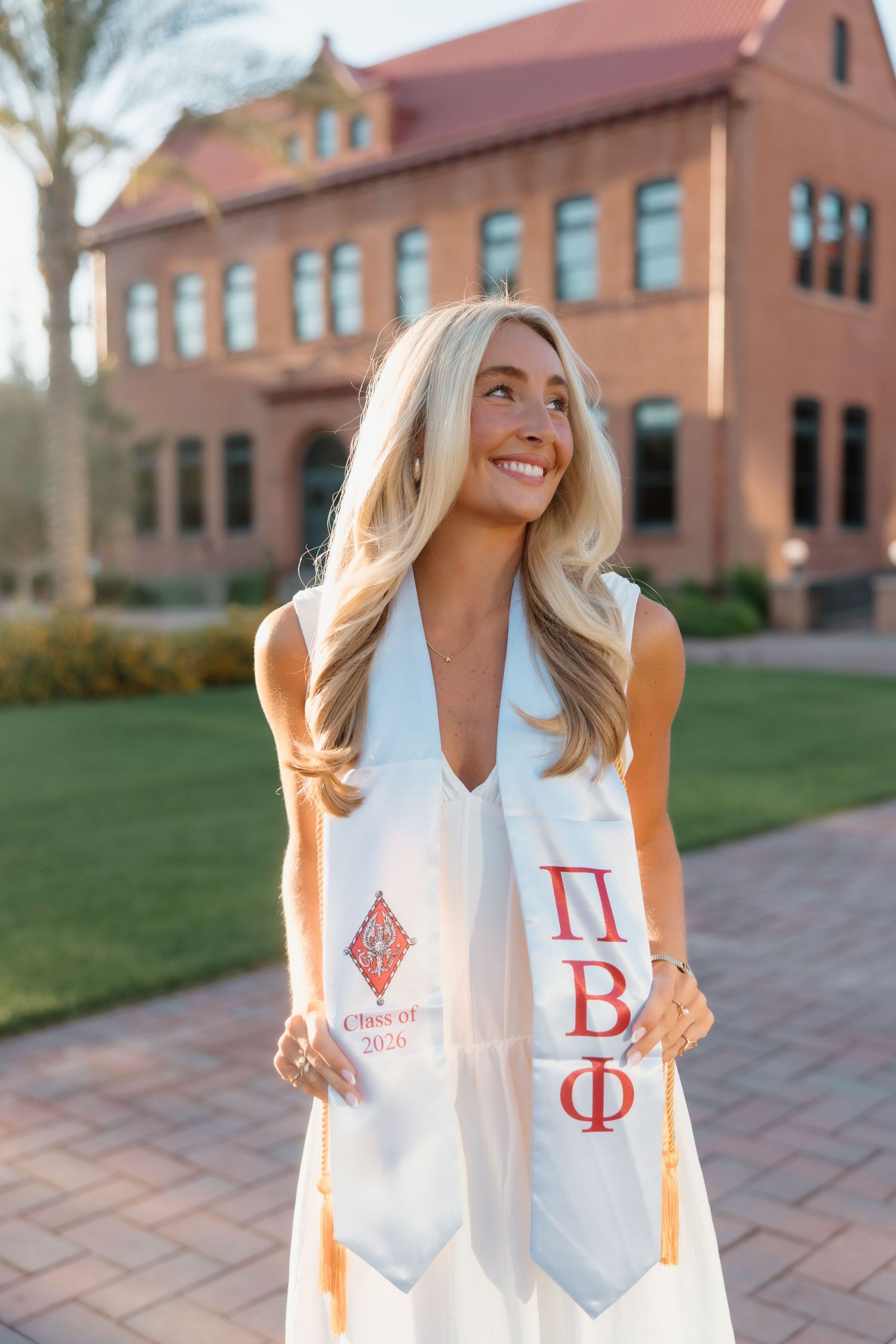 Blonde college graduate smiles outdoors wearing white dress and Pi Beta Phi graduation stole on sunny campus.