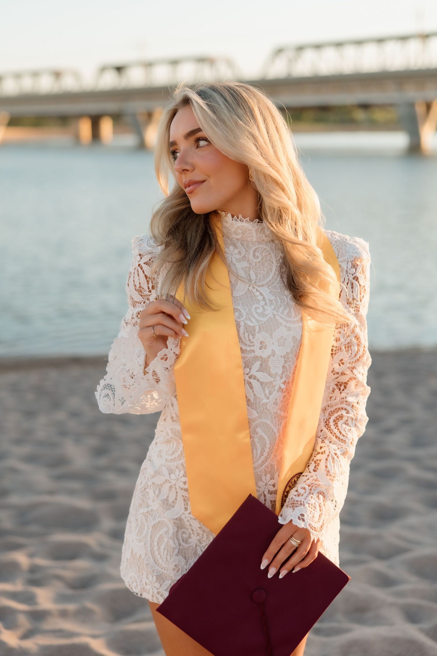 Blonde woman in white lace jacket and yellow dress holding burgundy clutch on sandy beach near bridge.