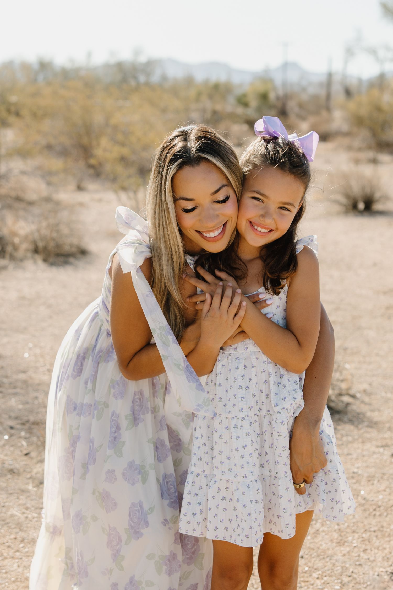 A smiling woman and young girl in matching floral dresses share a warm hug outdoors in a desert landscape.