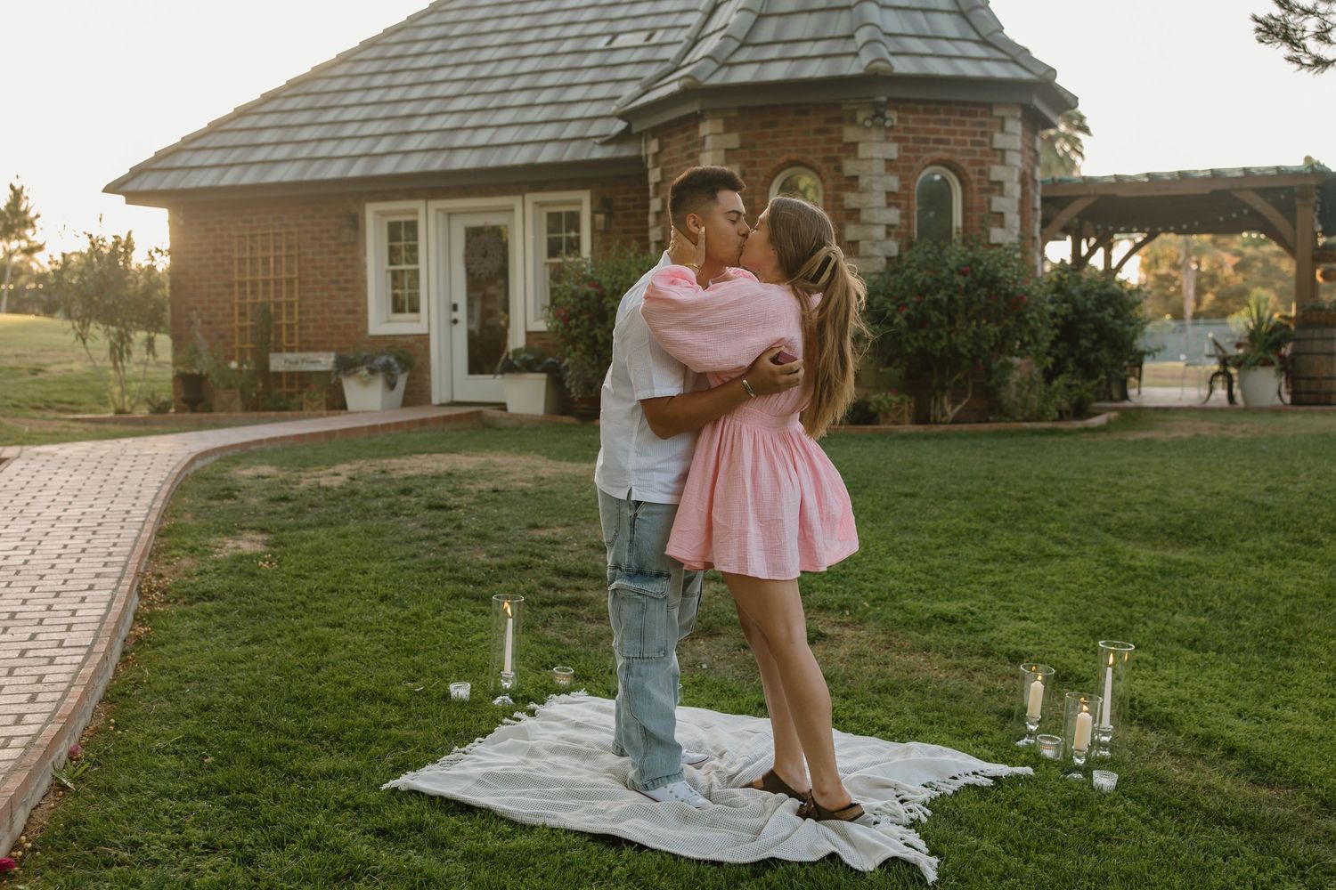 Romantic couple embracing on white blanket with candles in garden near brick cottage at sunset.