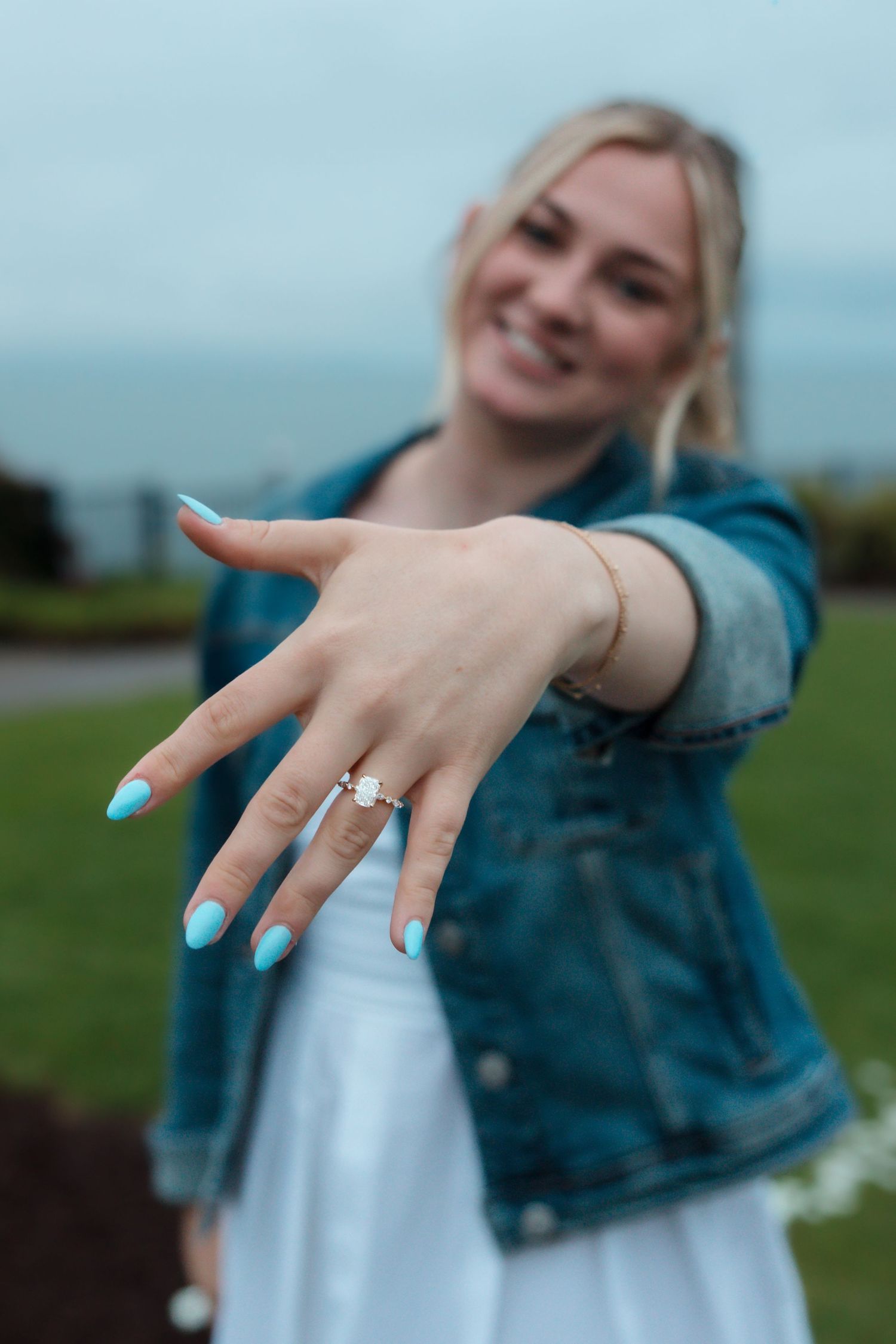 Smiling blonde woman showing off diamond engagement ring with teal nail polish outdoors.