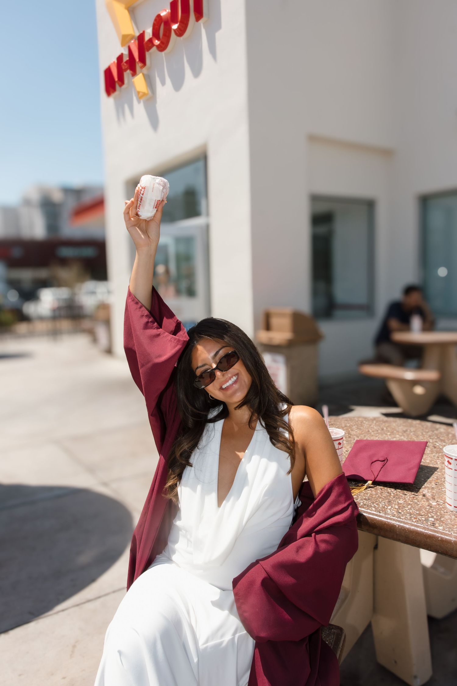 Smiling graduate in white dress and maroon gown raising a drink at In-N-Out Burger.