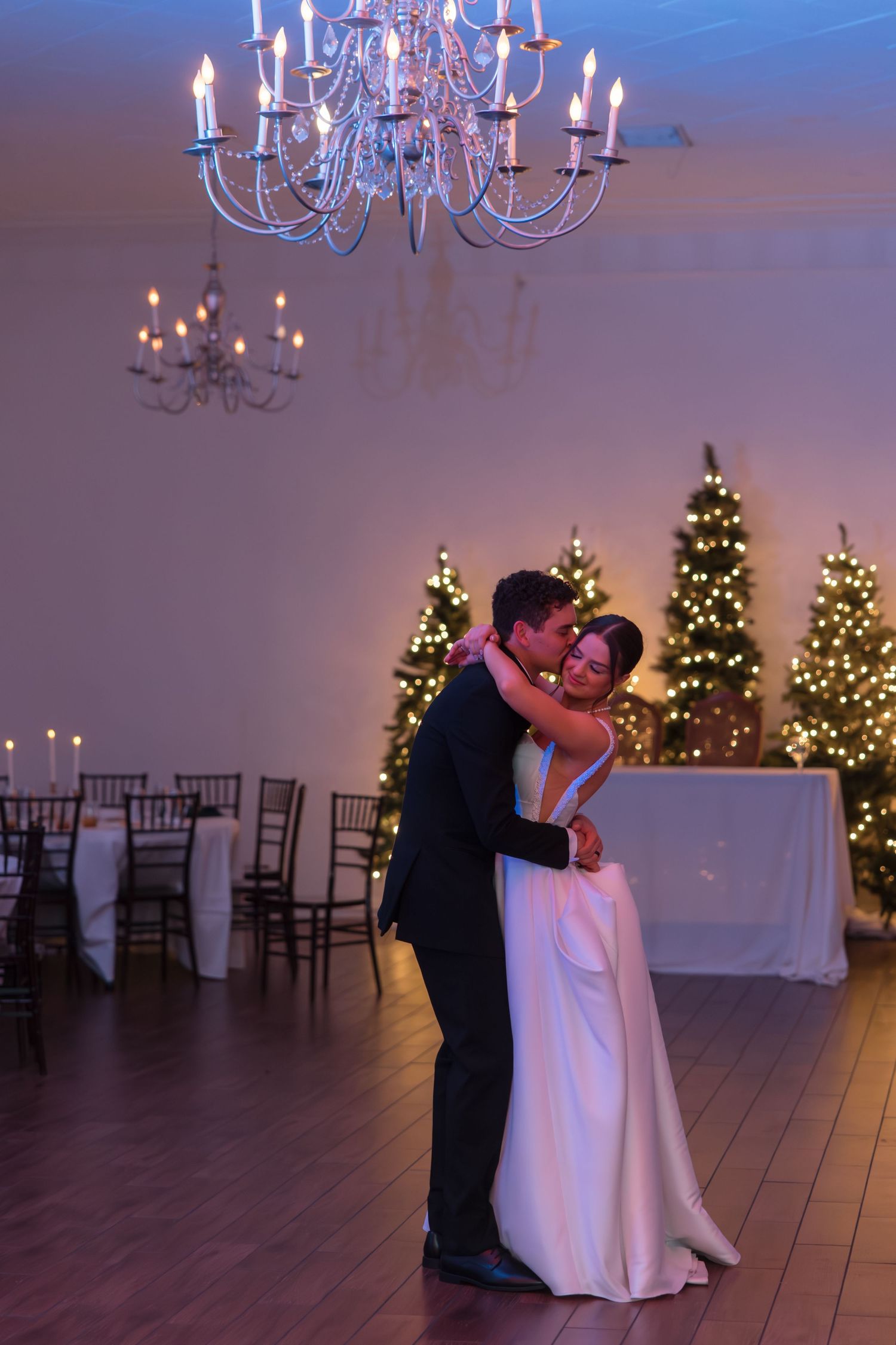 Couple embraces during first dance under crystal chandelier in softly lit wedding reception hall with Christmas trees.