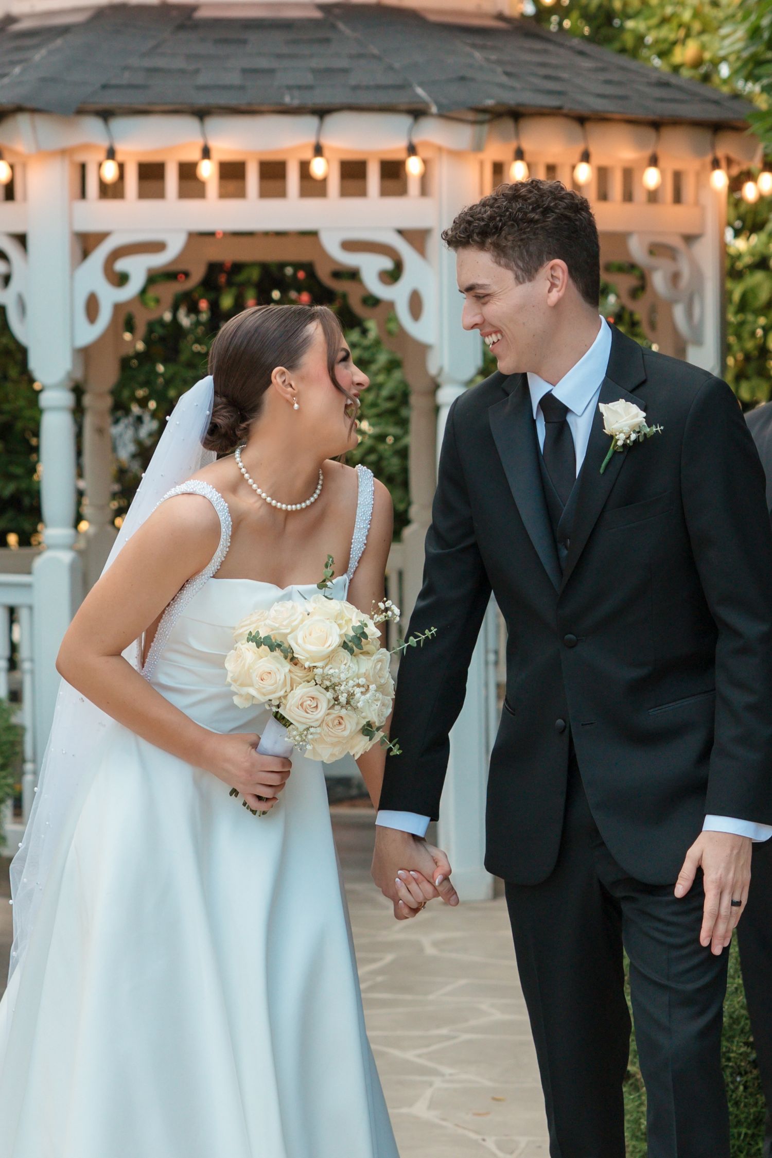 Happy bride and groom walking hand in hand near lit gazebo after wedding ceremony.