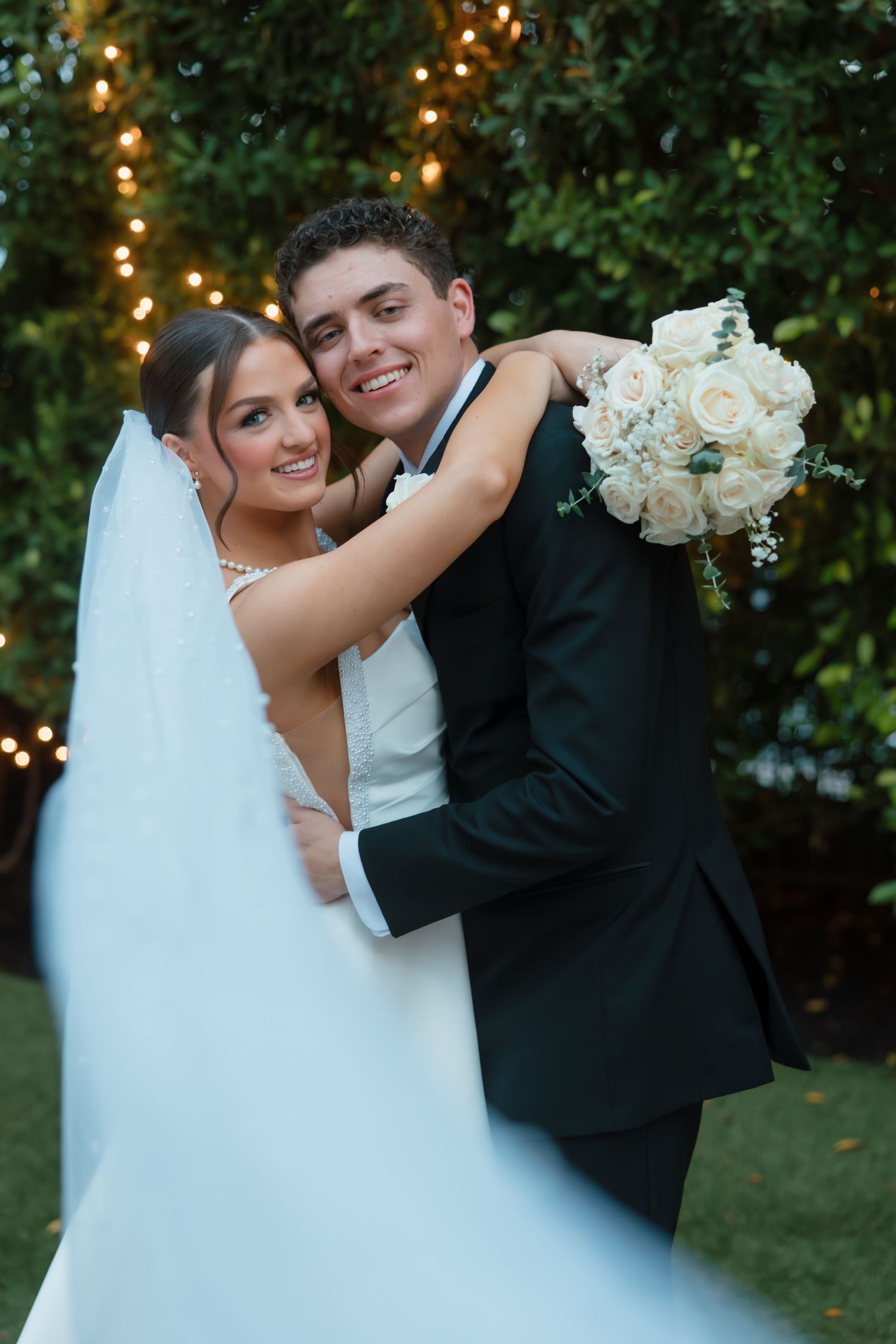 Smiling bride and groom embrace outdoors with white rose bouquet against a lighted hedge.