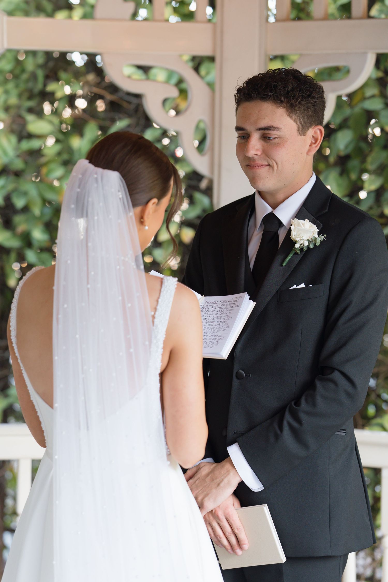 Groom in black suit reading handwritten vows to bride in white gown at outdoor wedding ceremony.