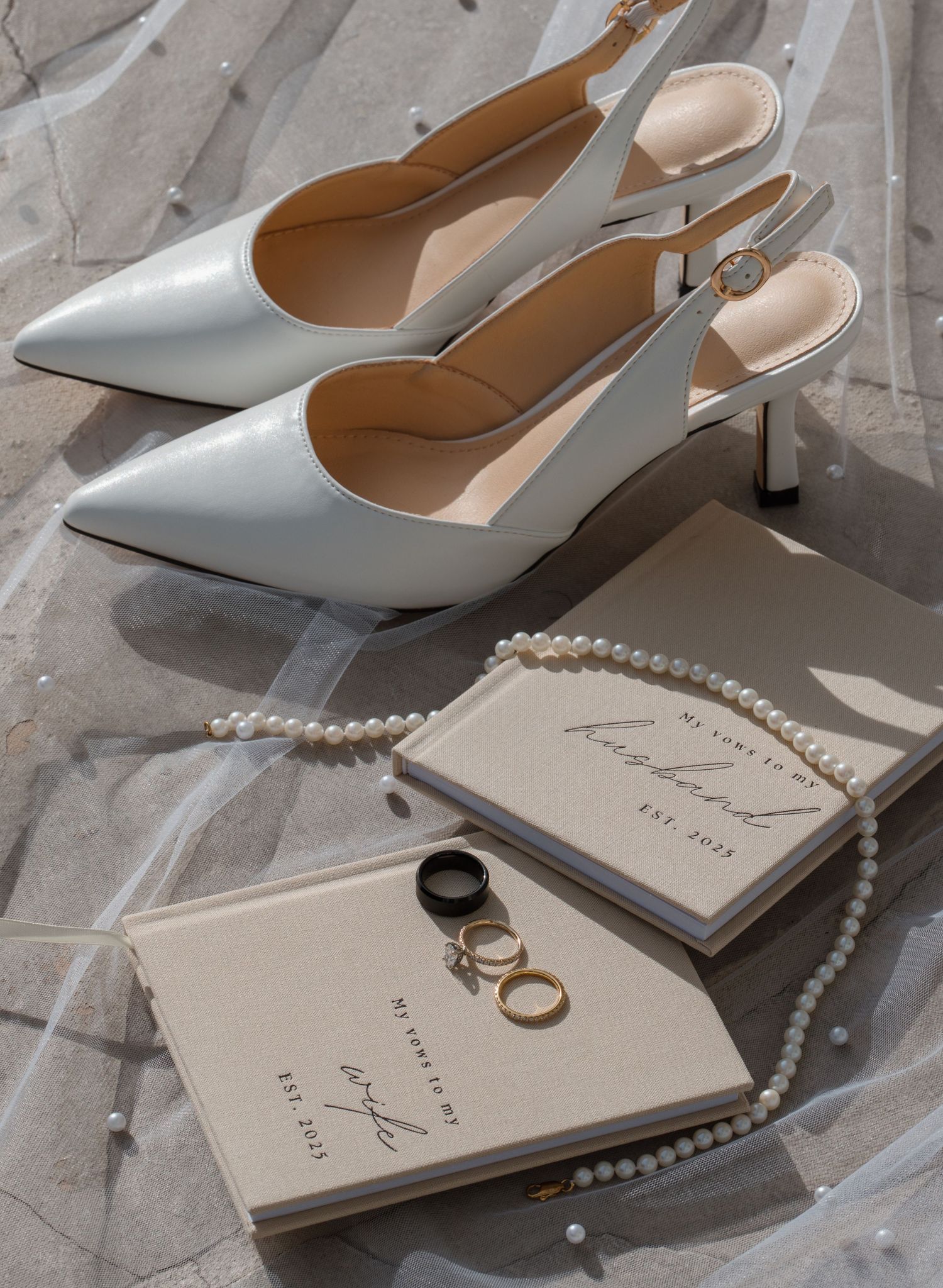 White slingback heels, vow books, pearl necklace, and wedding rings arranged on bridal veil.