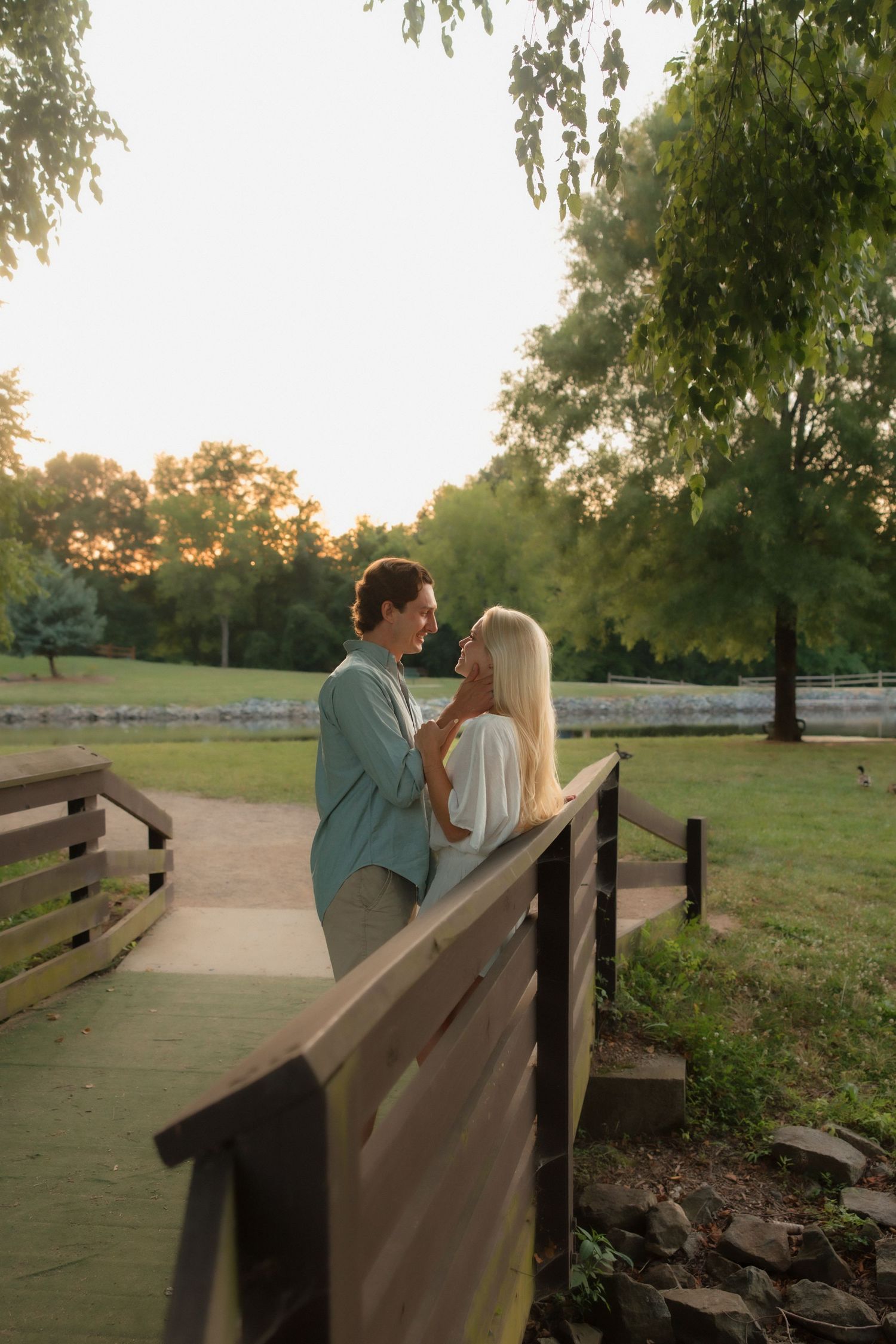 Young couple gazing into each others eyes on a wooden bridge surrounded by weeping willow trees at sunset.