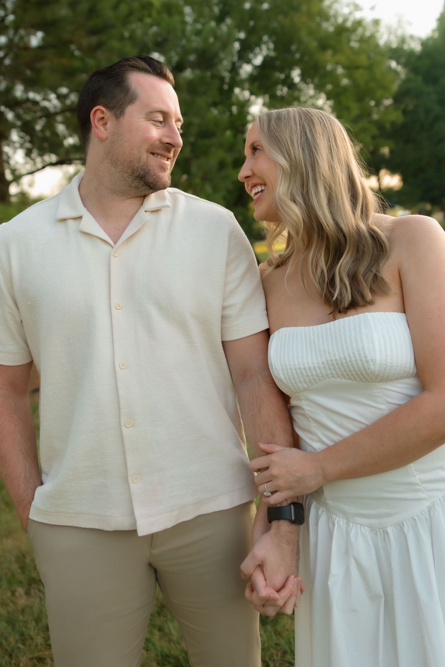 Happy couple holding hands and smiling at each other outdoors in park during engagement photo session.