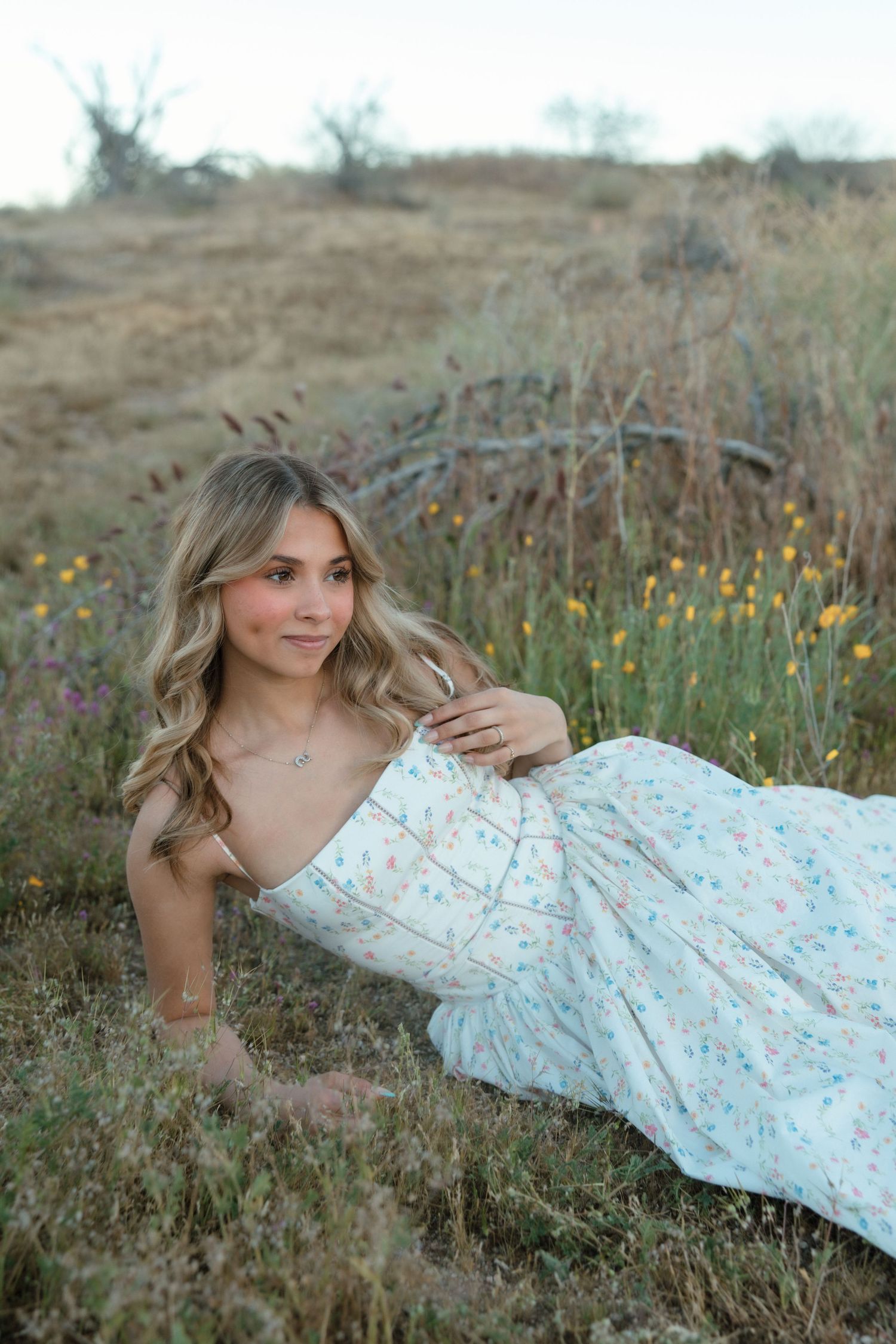 Young blonde woman in white floral dress reclining in natural wildflower meadow for senior photos.