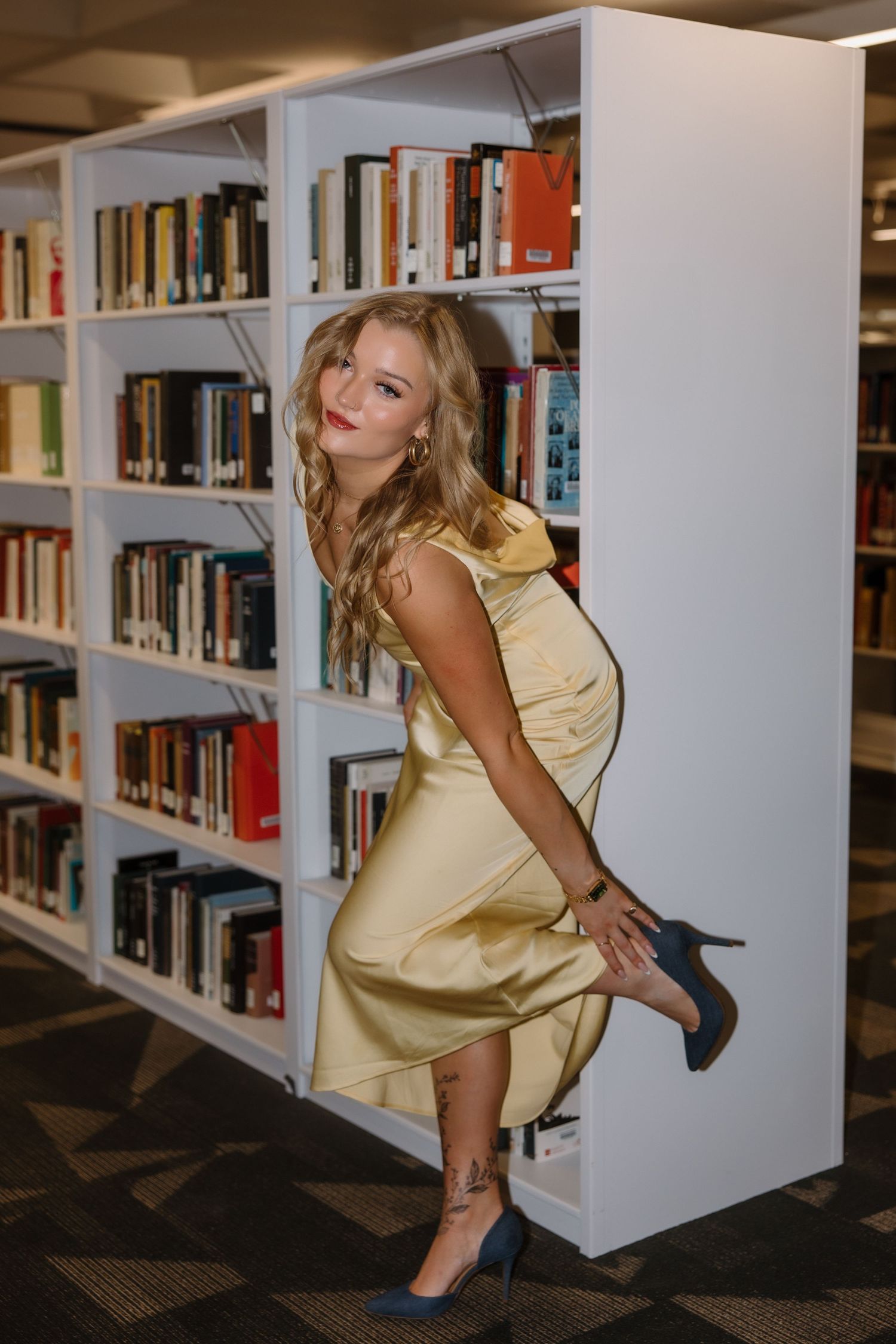Young woman in gold satin dress posing elegantly beside white library bookshelves with heels.