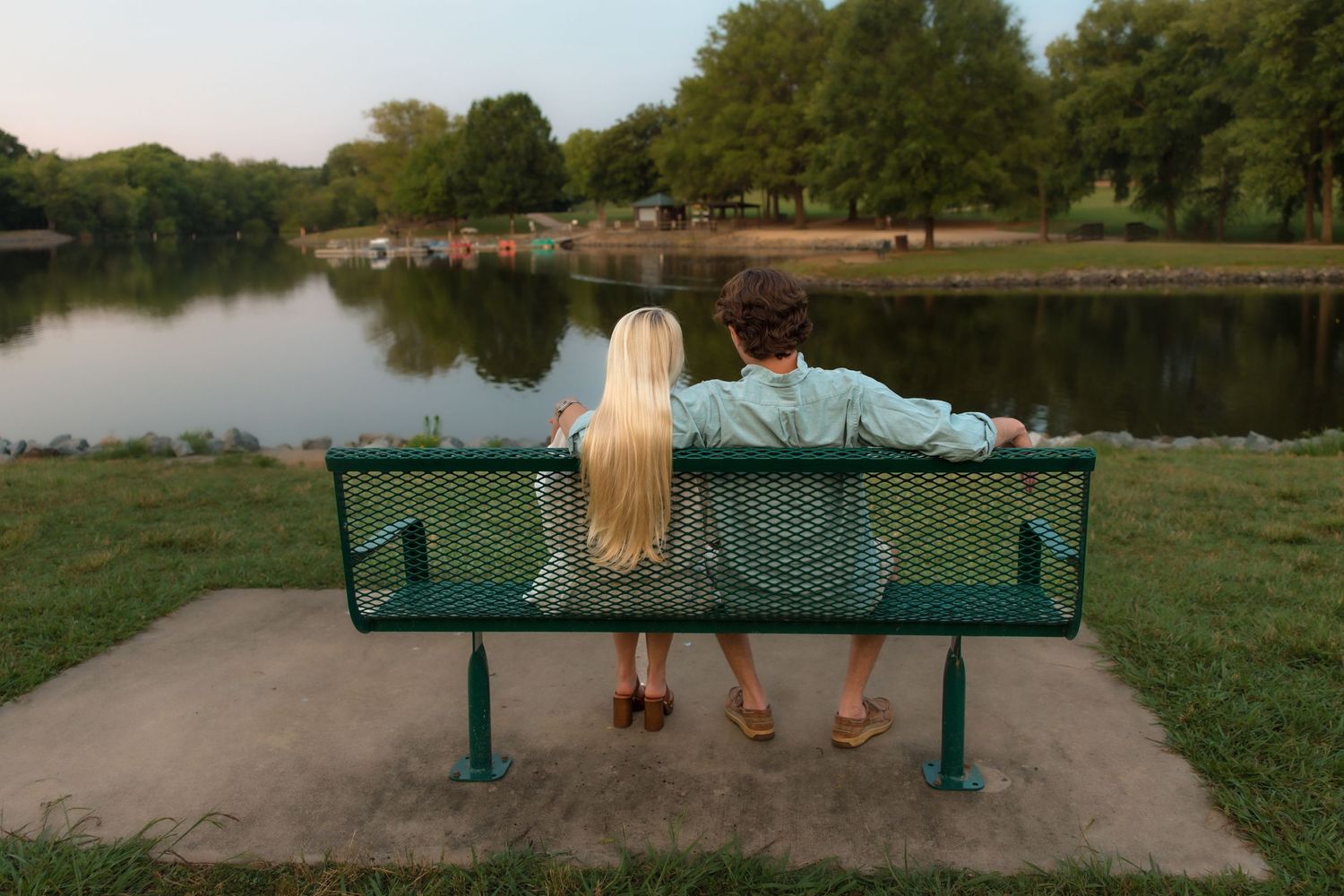 Couple sitting together on a green bench overlooking a calm lake surrounded by trees at sunset.