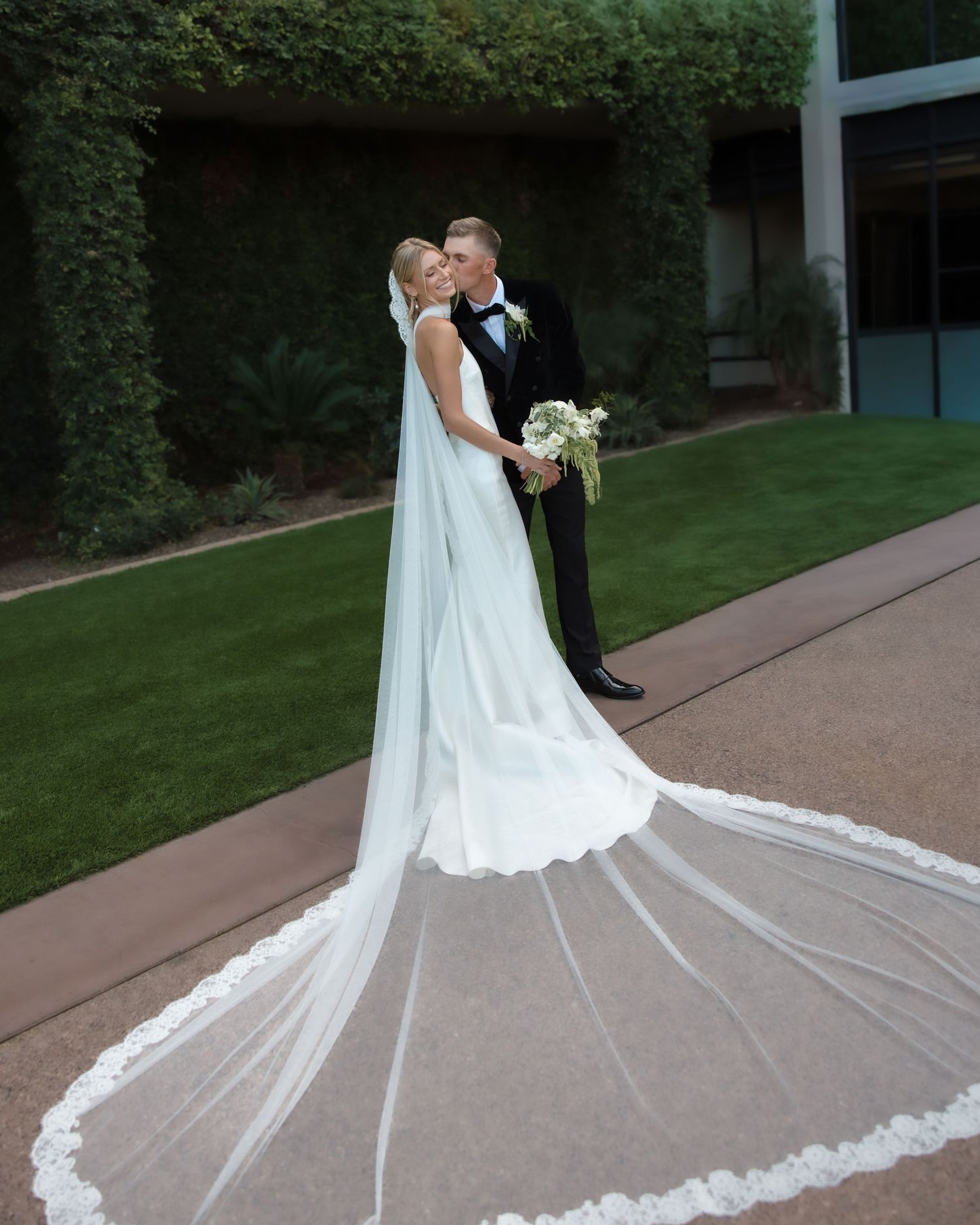 Bride in white gown with lace cathedral veil kisses groom in black tuxedo in garden setting.