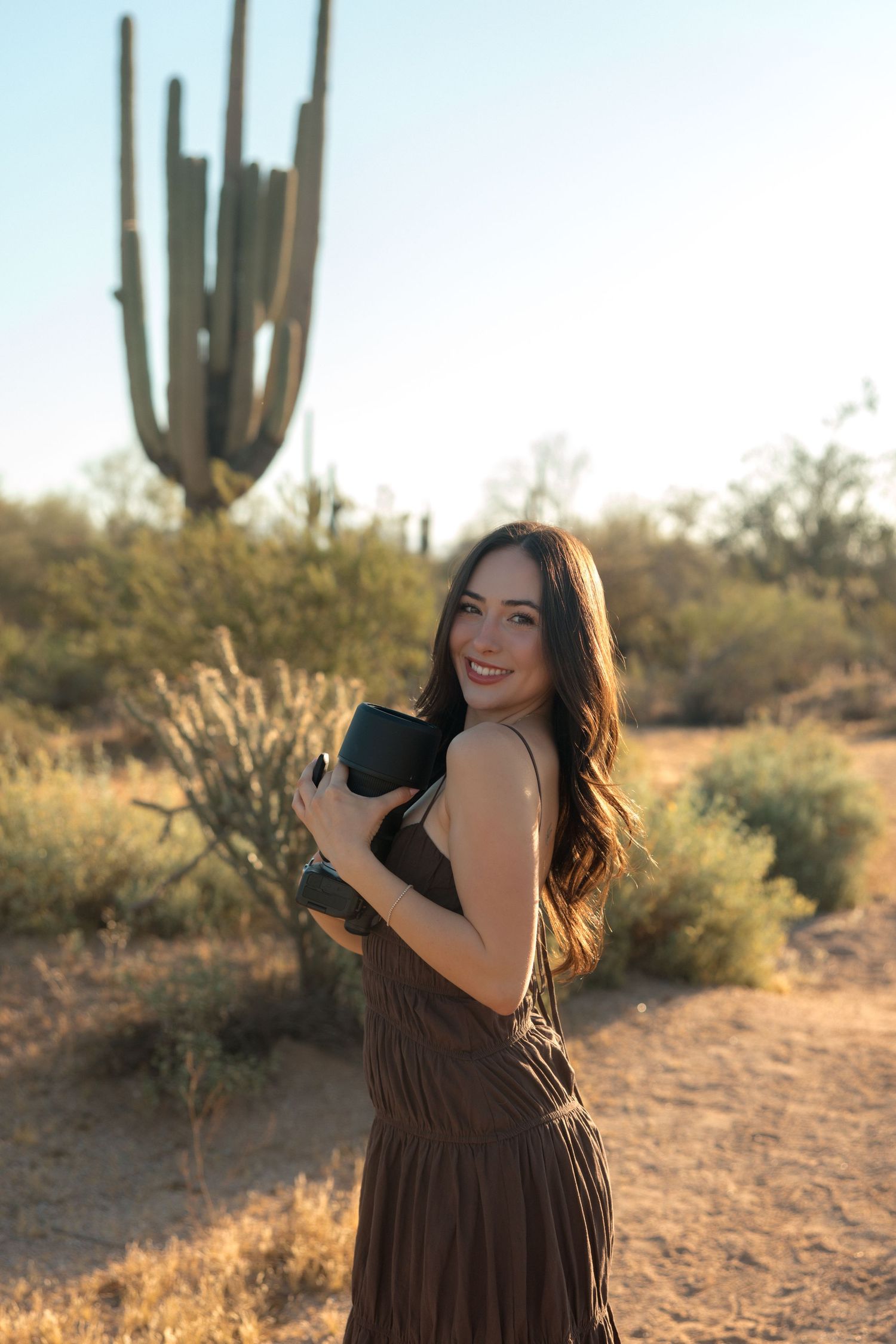Young woman in brown sundress holds camera and smiles in Arizona desert with tall saguaro cactus.