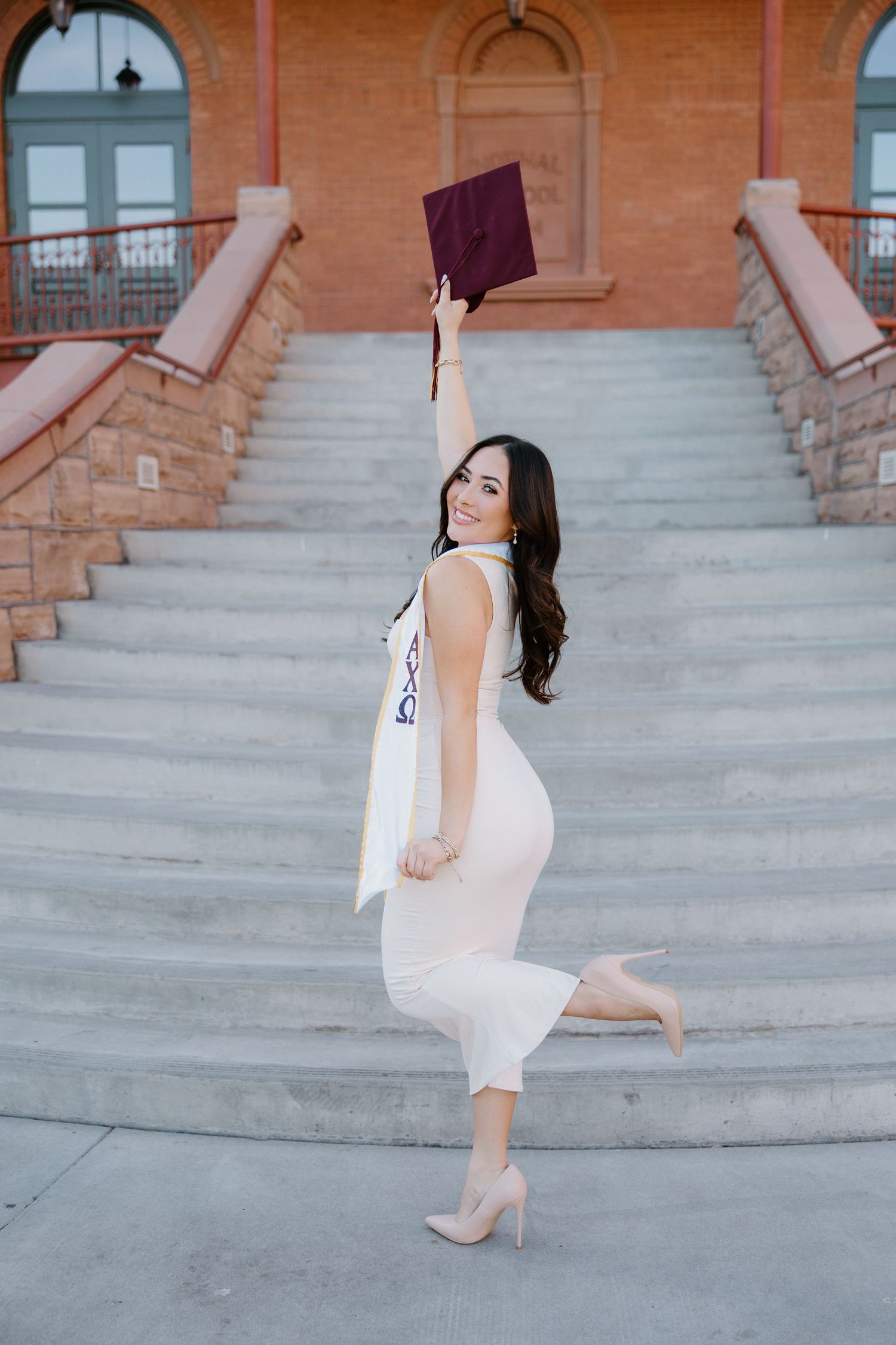Joyful graduate in white dress raising maroon cap on steps outside historic brick building.