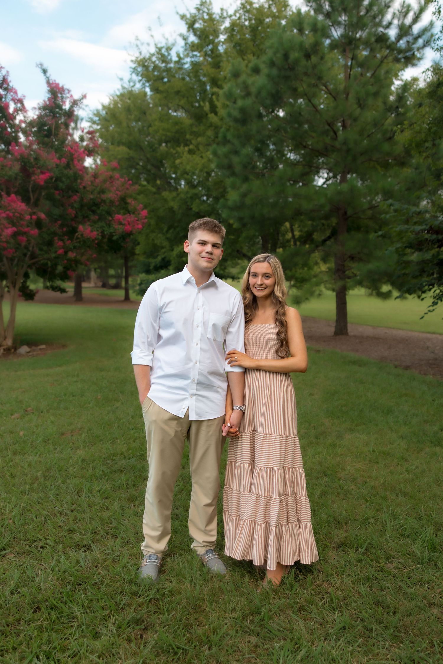 Young couple holding hands in a lush green park, man in white shirt and khakis, woman in a flowing blush maxi dress.