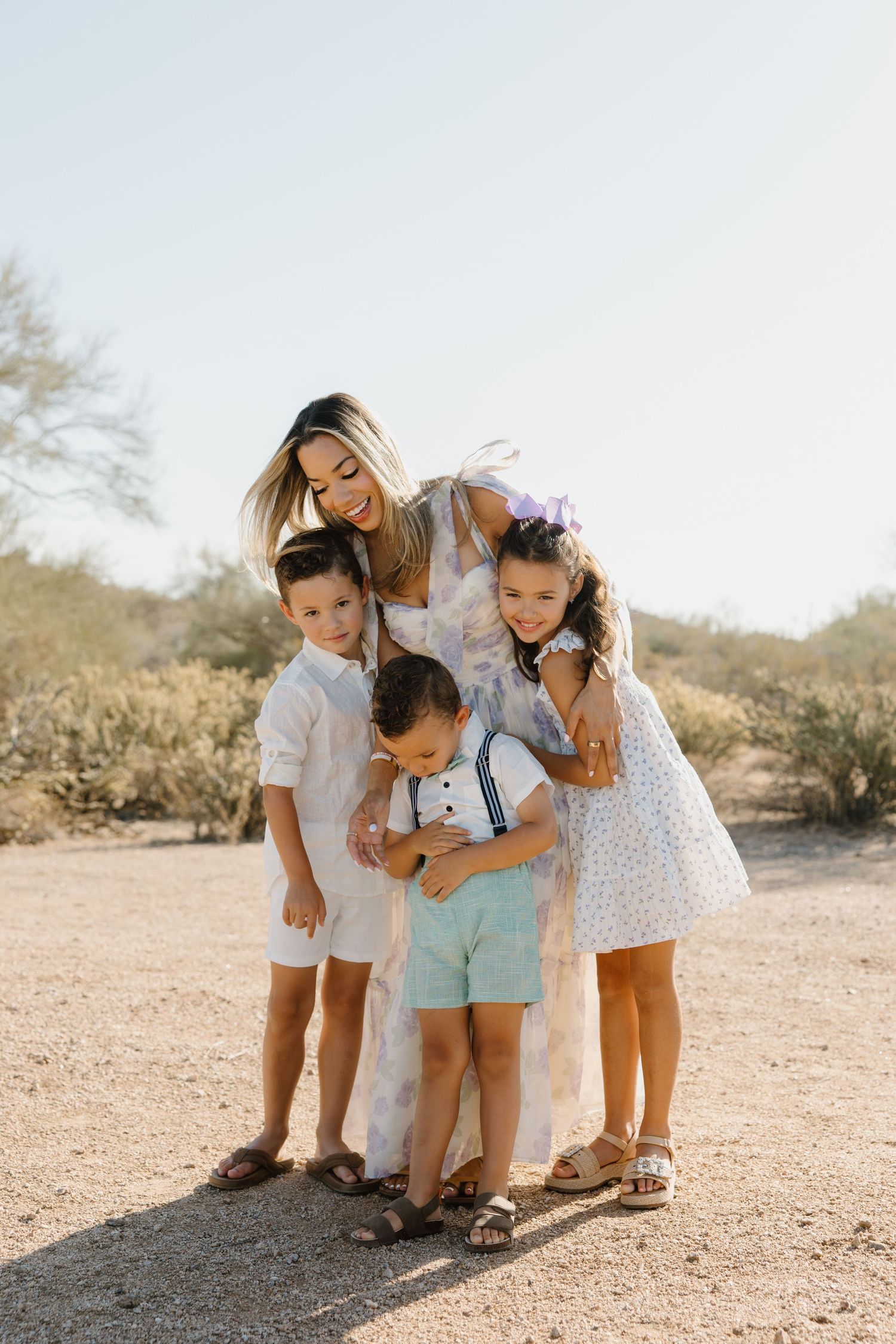 A woman smiles while hugging three young children outdoors in a desert landscape, all dressed in light, coordinated outfits.