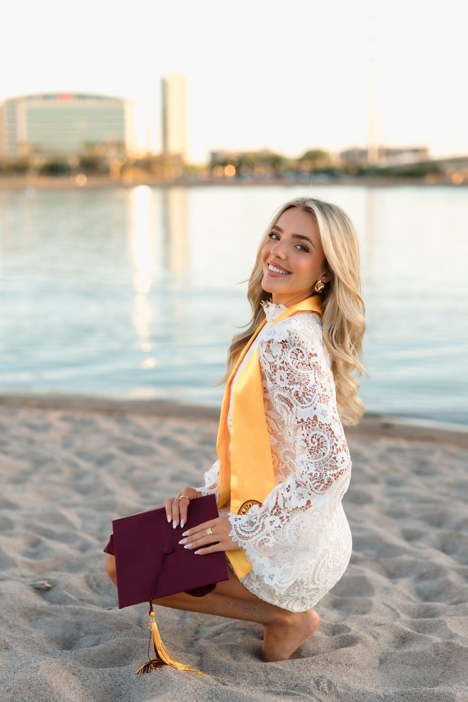 Smiling blonde graduate in white lace dress holds maroon cap while kneeling on sandy beach.