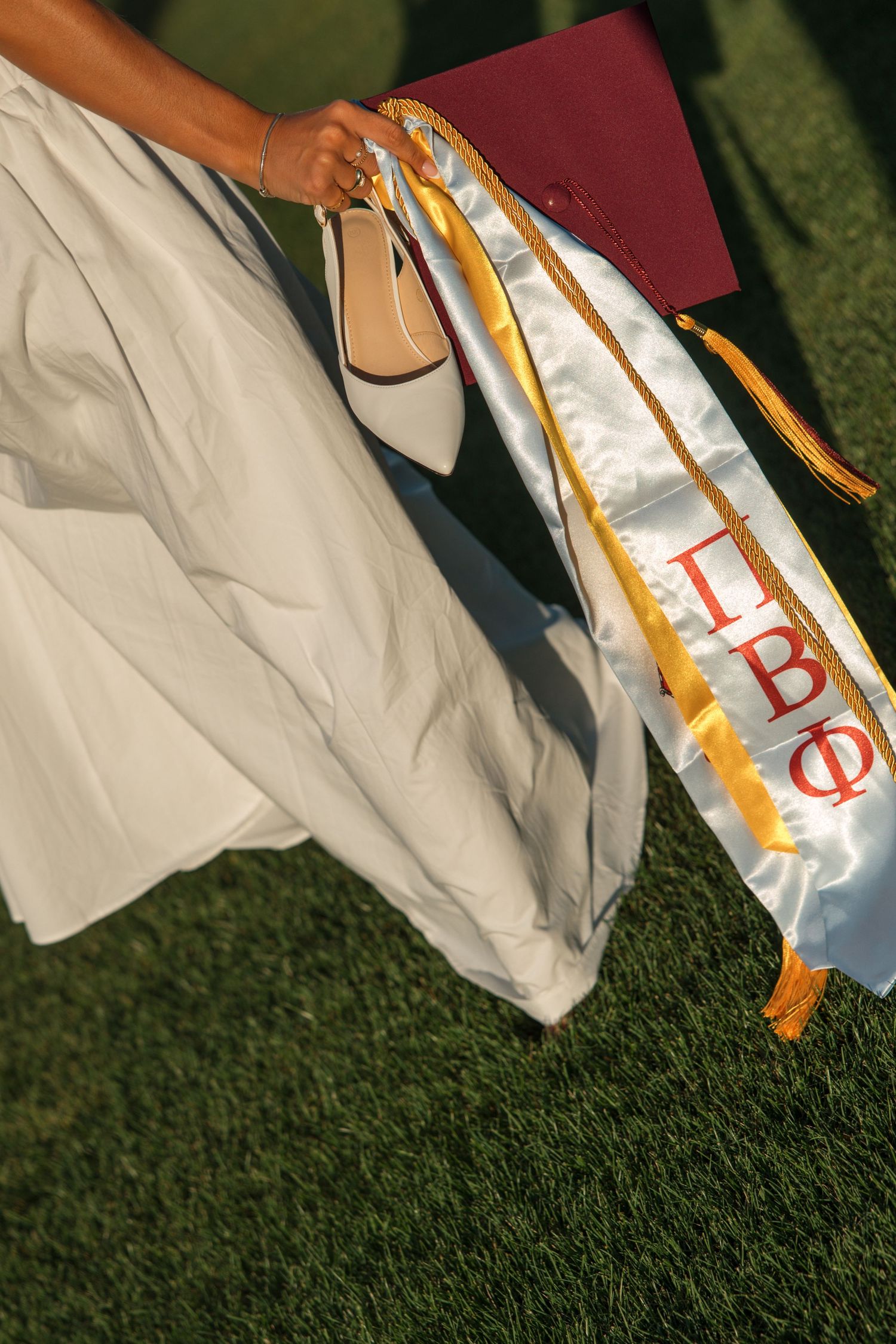 Close-up of Pi Beta Phi graduation stole, maroon cap, white heels and gown held on green grass.