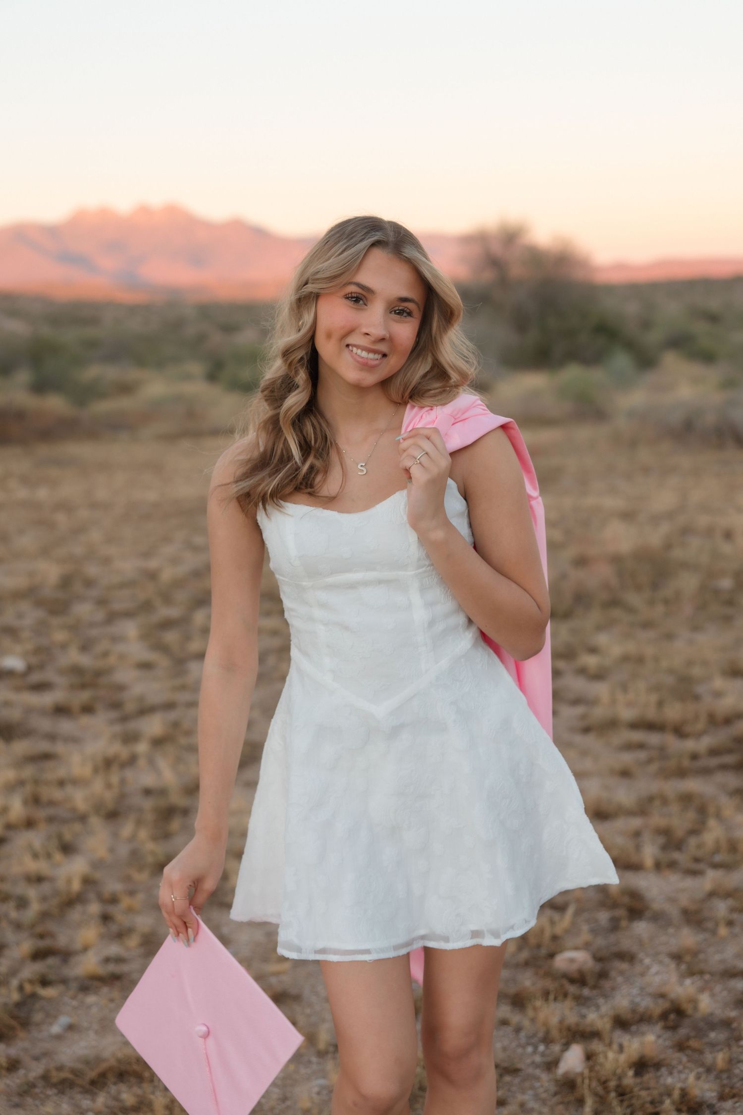 Young woman in white dress holding pink graduation cap in desert landscape at sunset.