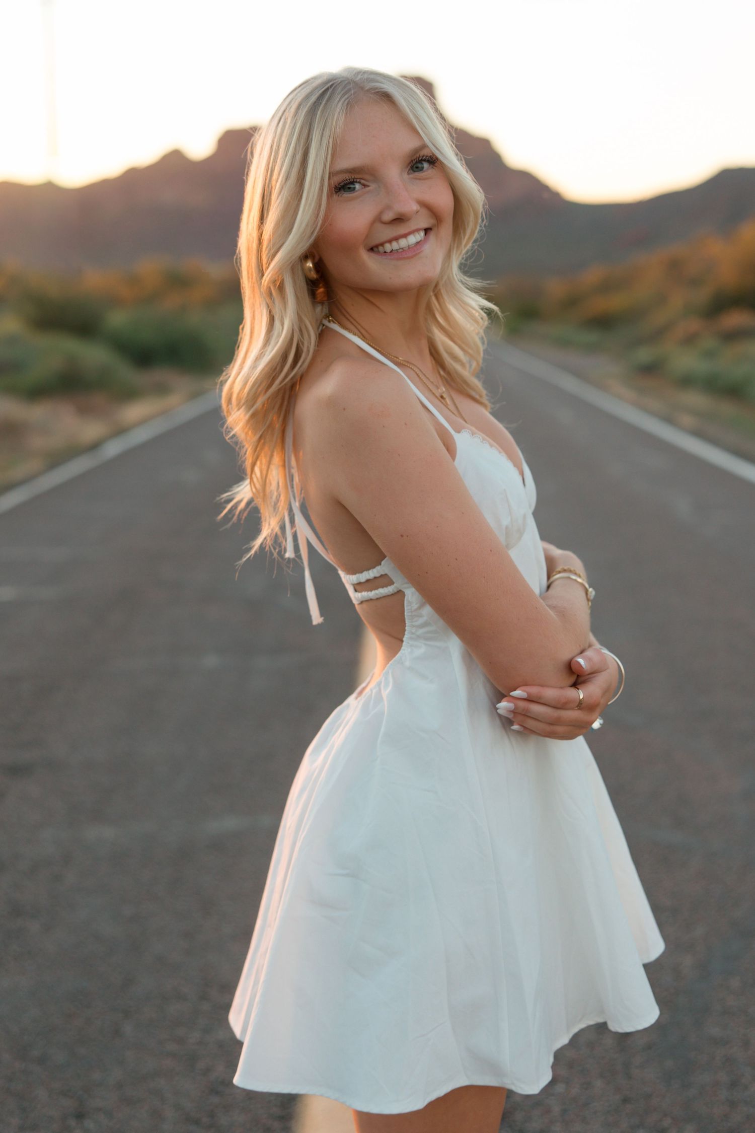 Smiling blonde woman in white sundress standing on desert road at sunset with mountains in background.