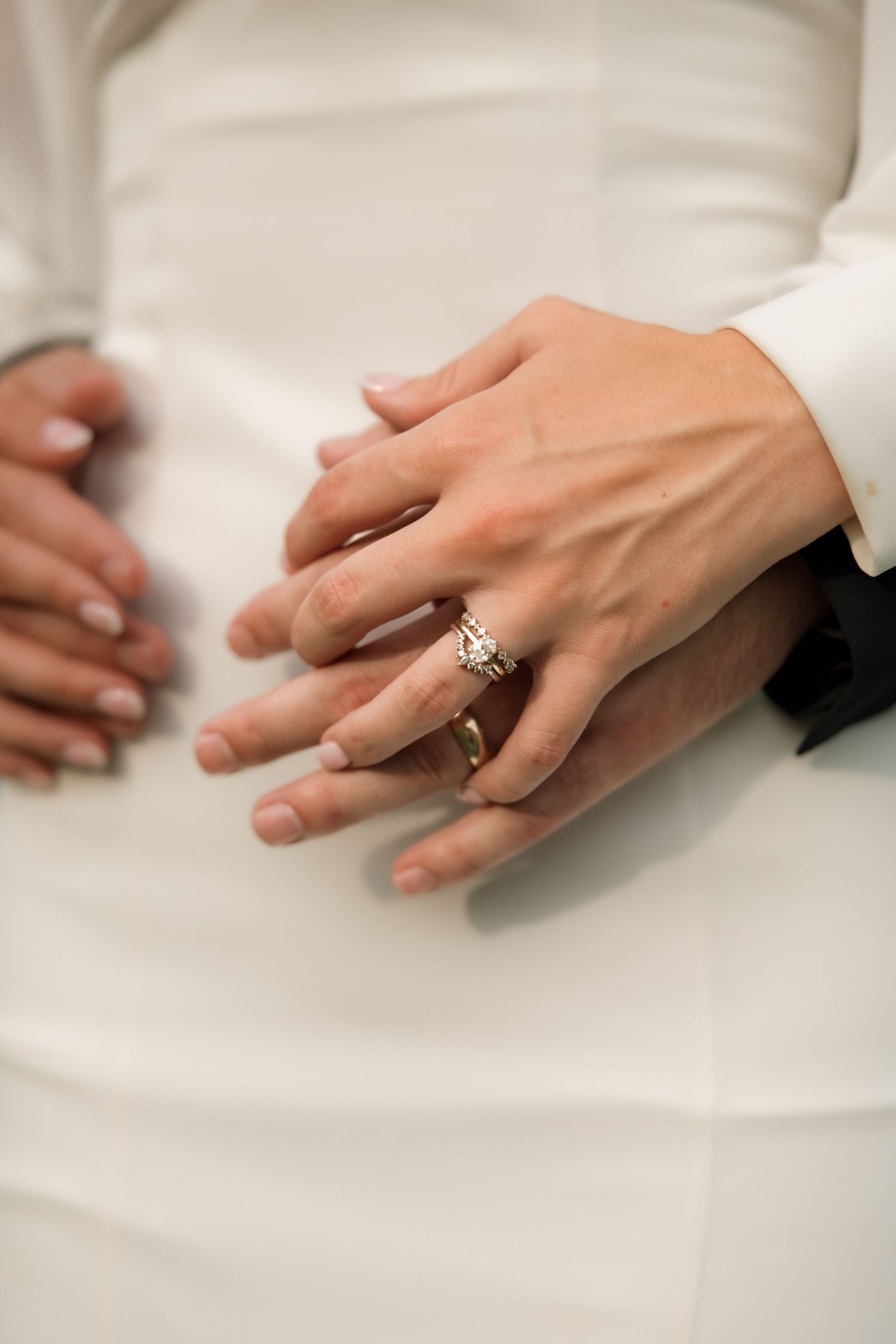 Close-up of bride and groom hands showing diamond engagement ring and wedding bands on white gown.
