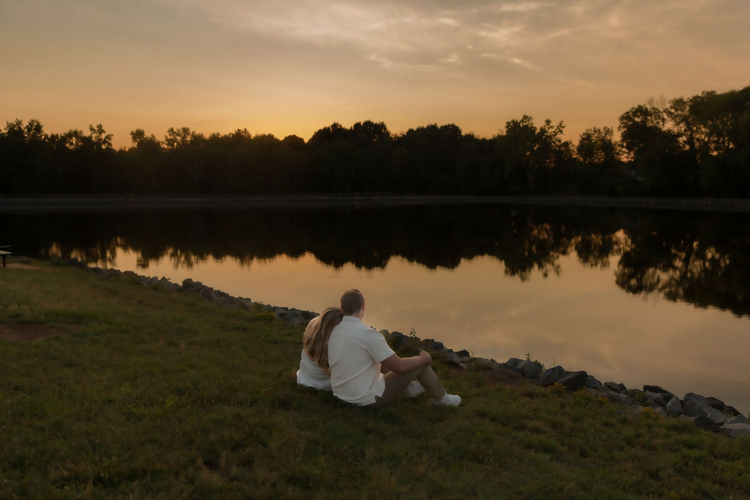 Couple sitting together on grass watching a stunning golden sunset reflect across a peaceful lake.