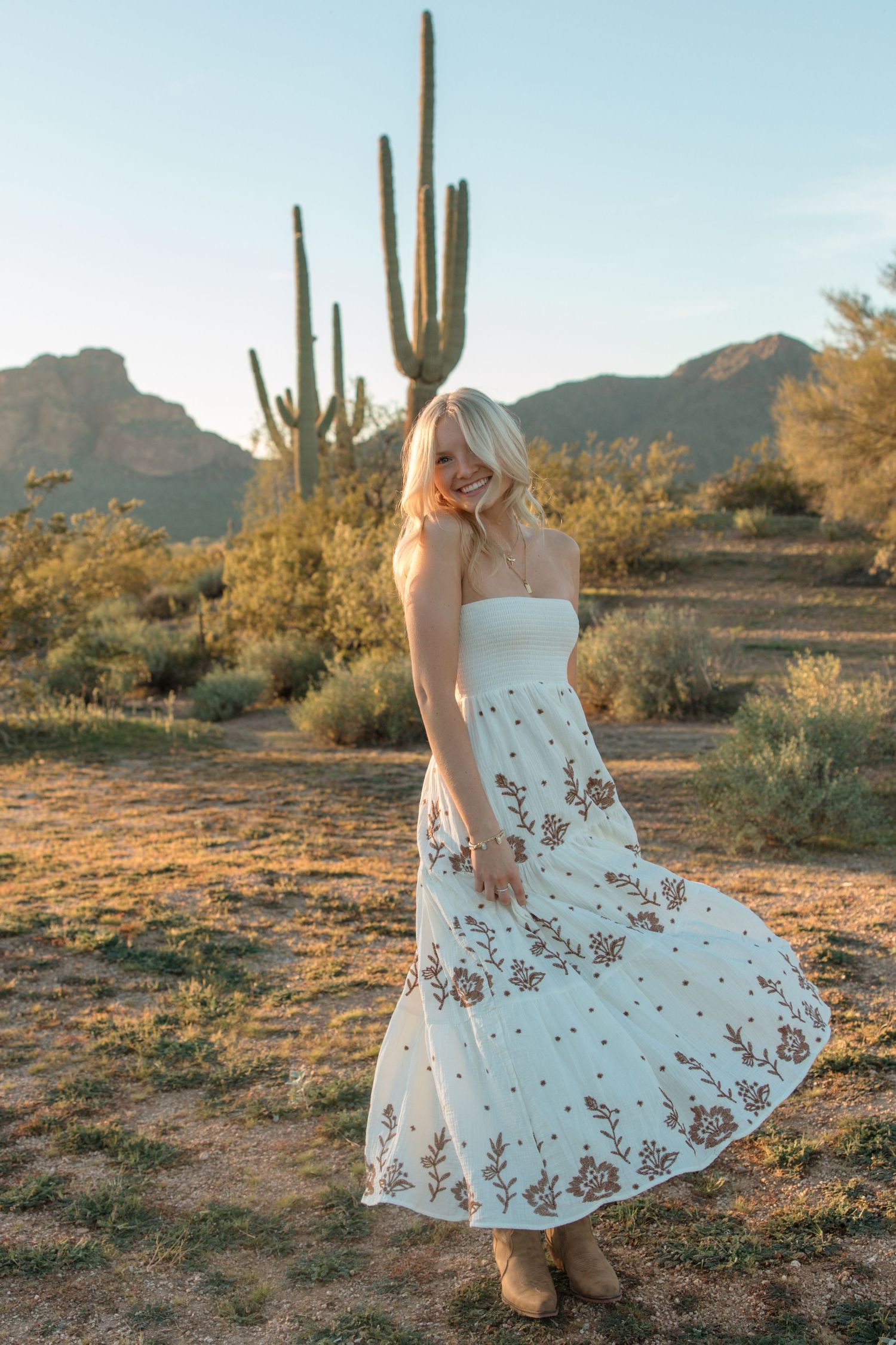 Smiling blonde woman twirling white embroidered maxi dress in Arizona desert with saguaro cactus at sunset.